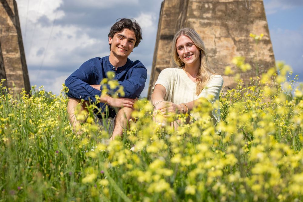 Two people sit together in a field of yellow wildflowers with stone architecture in the background.