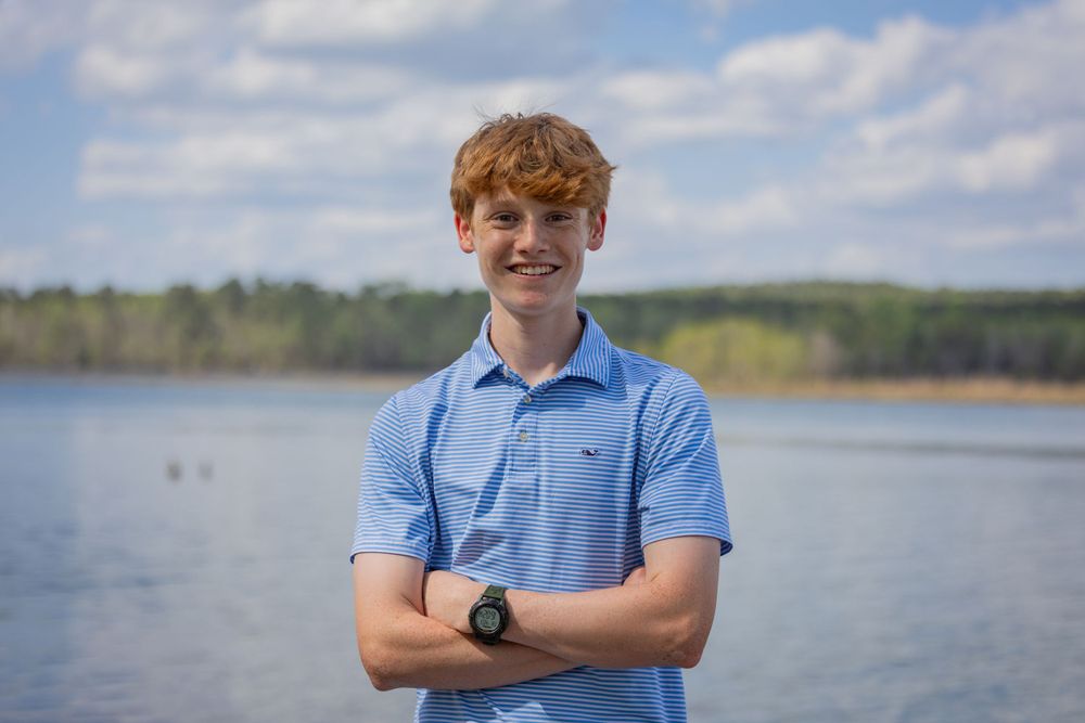 A person in a striped polo shirt poses with crossed arms by a lakeside.