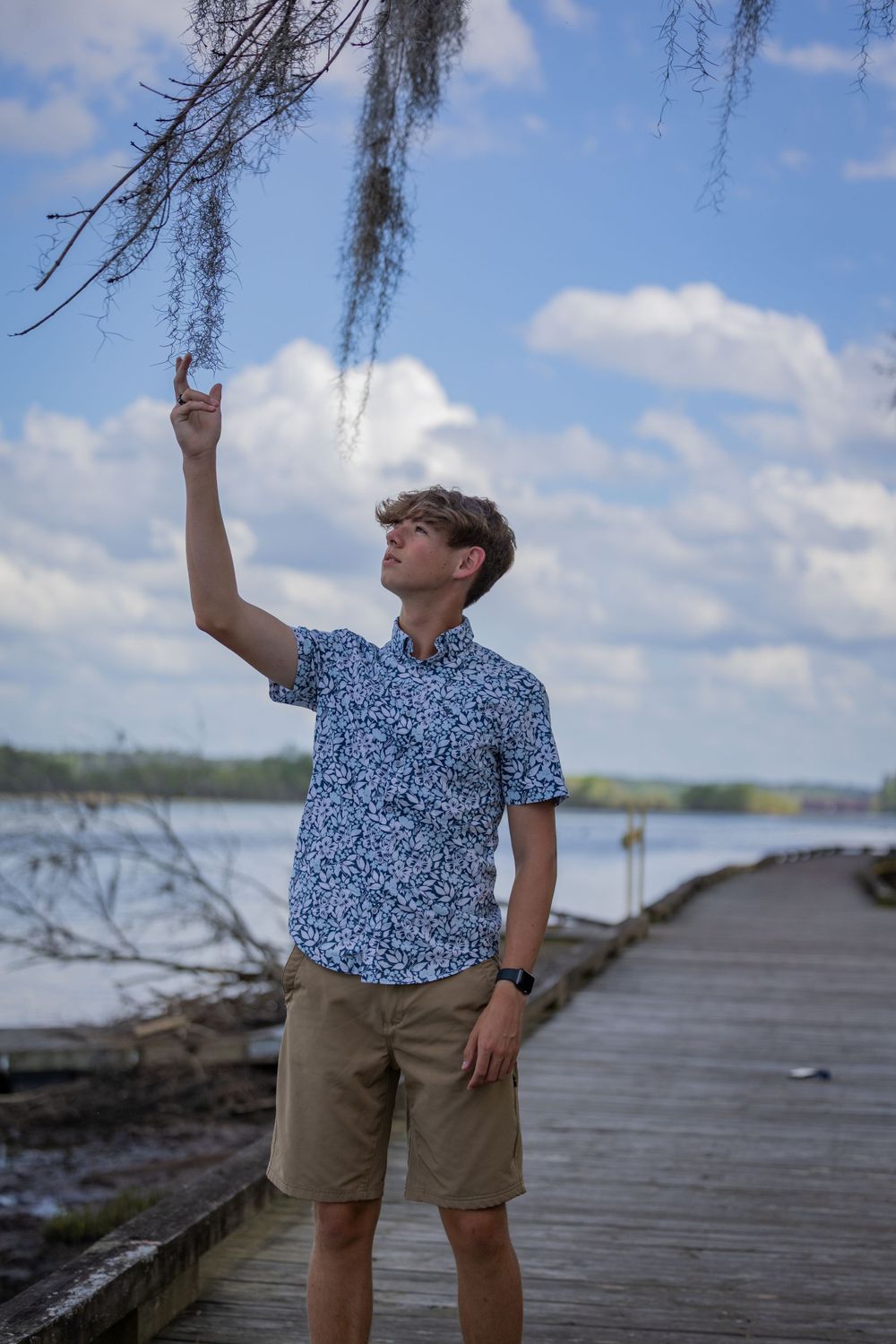 Person in blue patterned shirt and khaki shorts stands on boardwalk touching Spanish moss against cloudy sky.