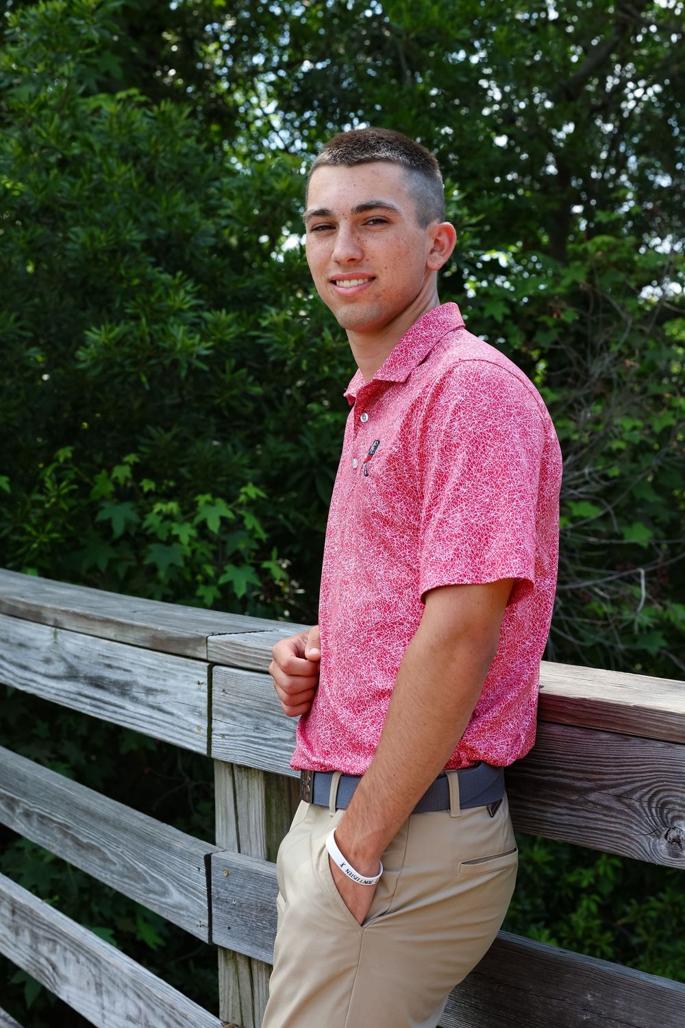 A person wearing a pink patterned button-down shirt and khaki pants poses outdoors by a wooden railing.