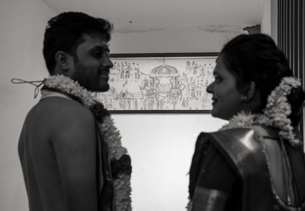 A black and white photograph shows two individuals wearing flower garlands in conversation while facing each other.