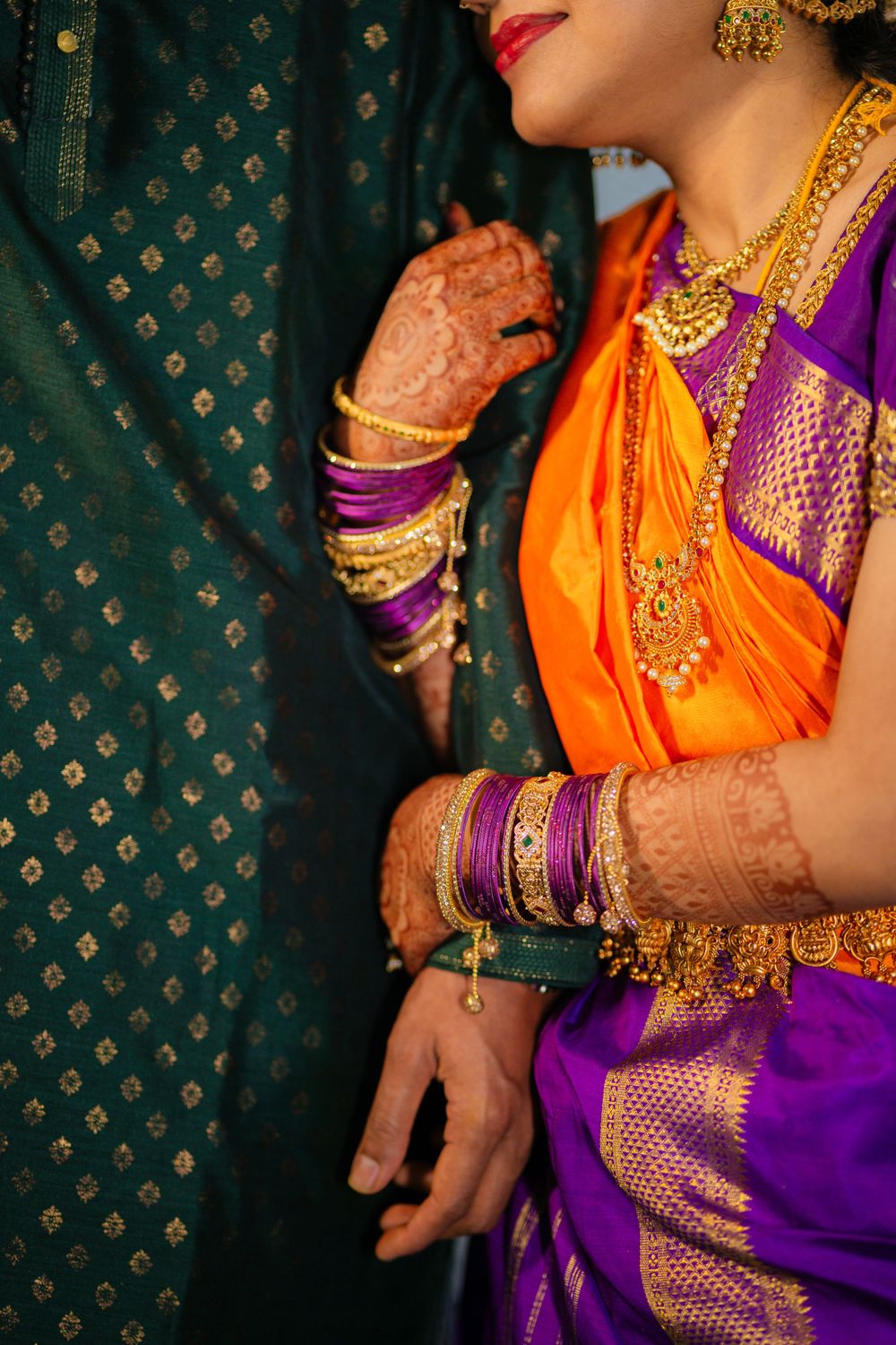 Close-up of traditional Indian bridal attire showing orange and purple silk sari with gold jewelry and henna decorated hands.