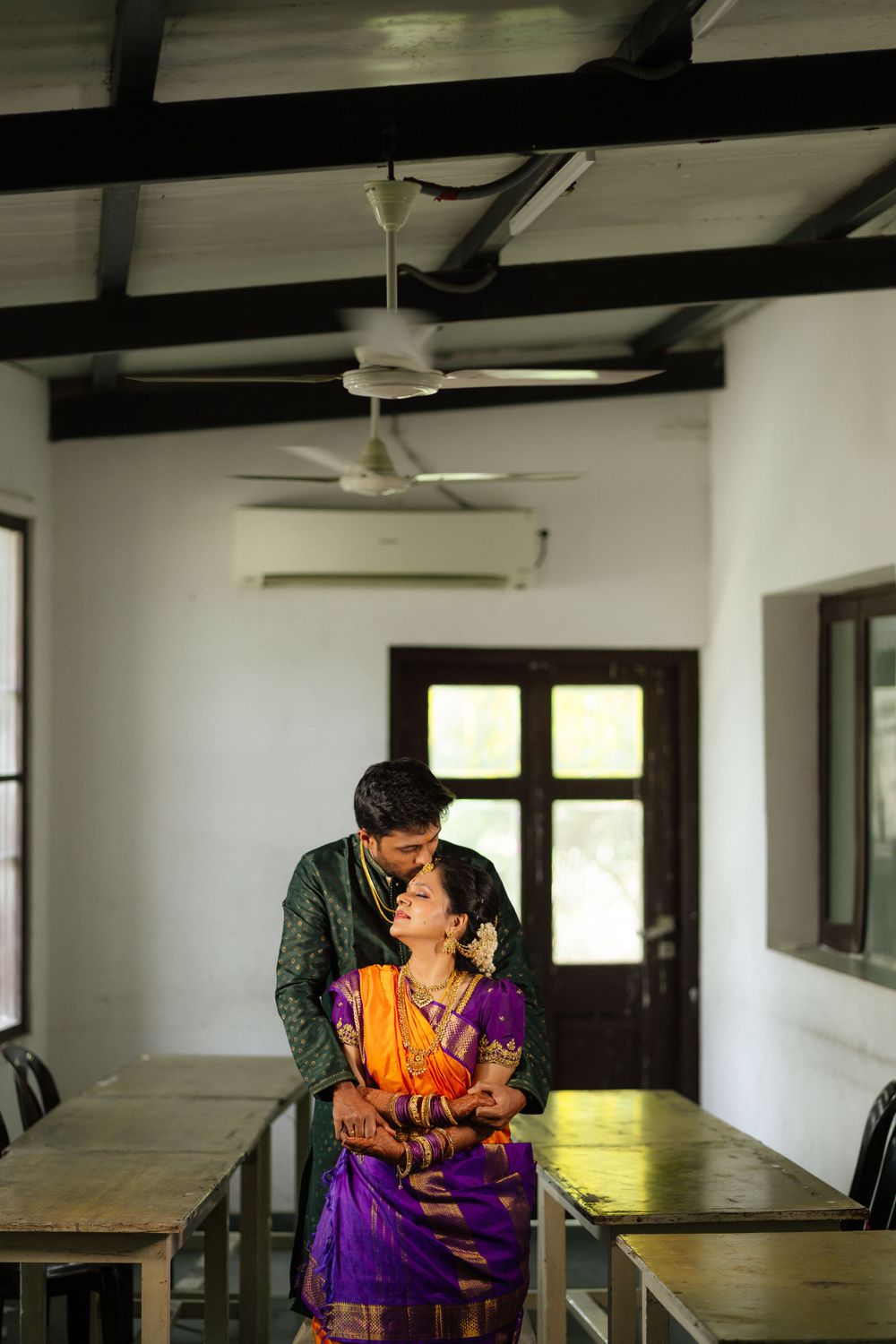 A couple in traditional Indian attire share a tender moment in a sunlit room with exposed wooden beams and desks.