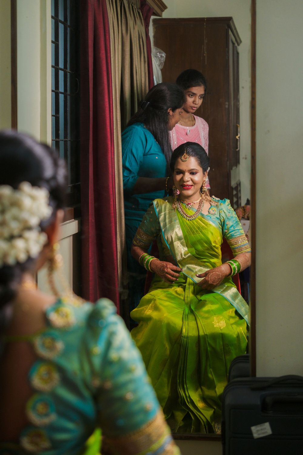 A person in a vibrant lime green silk sari with turquoise embellishments sits while getting ready in a mirrored room.