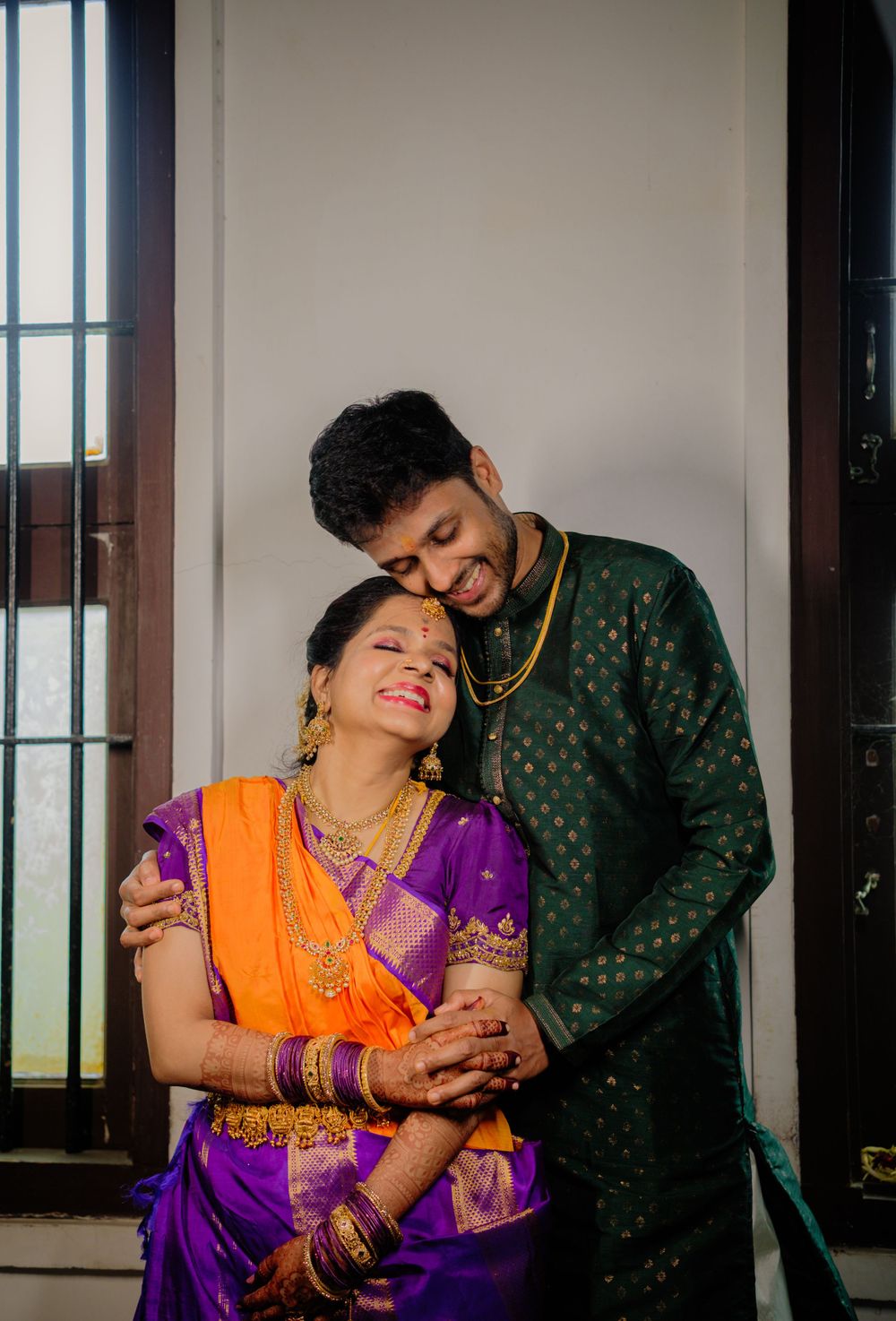 A couple in traditional Indian attire, wearing purple and green, share an intimate moment by a window.