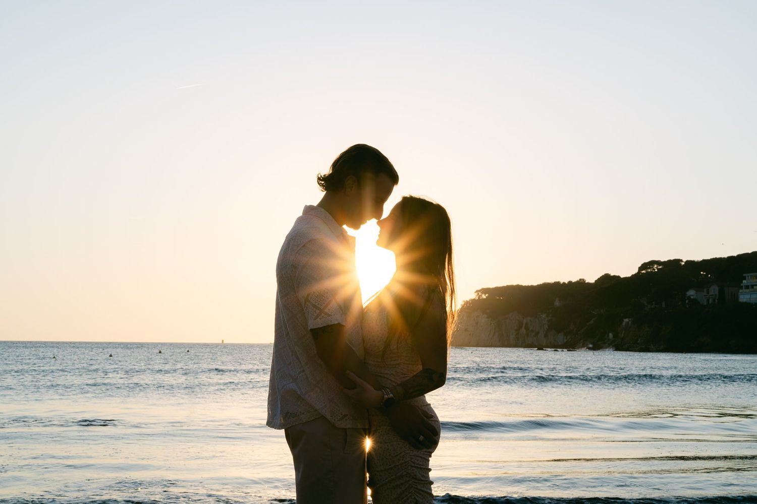 A silhouetted couple embraces on the beach at sunset with sunlight creating a starburst between them.