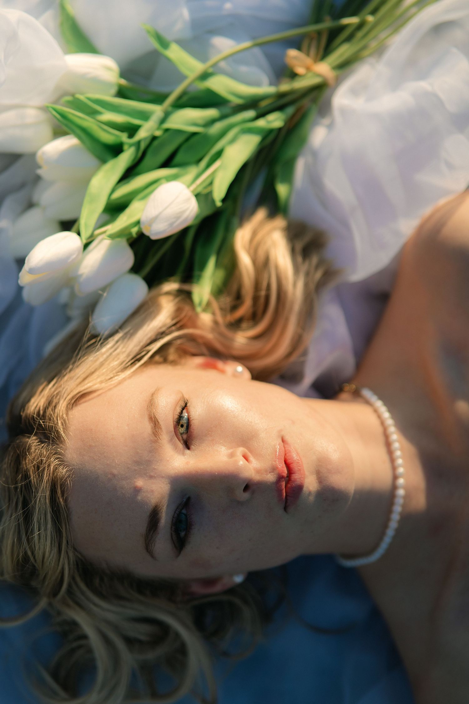 A soft portrait with white tulips and pearls laying against blue fabric in warm lighting.