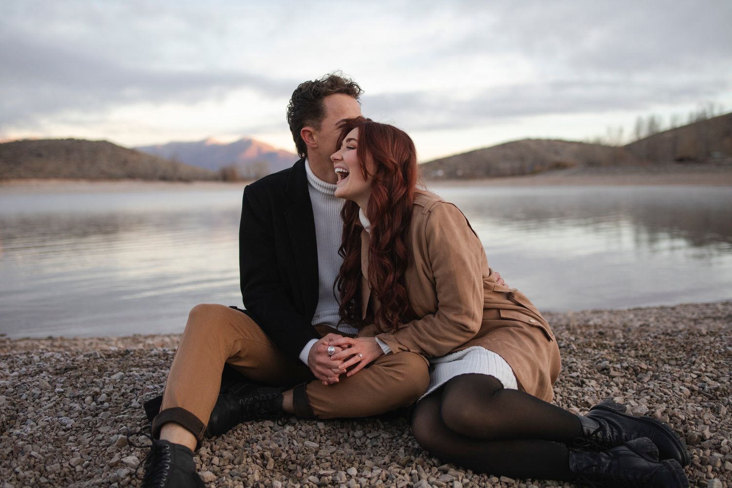 Couple laughs hilariously as they sit on the shoreline of a lake surrounded by mountains.