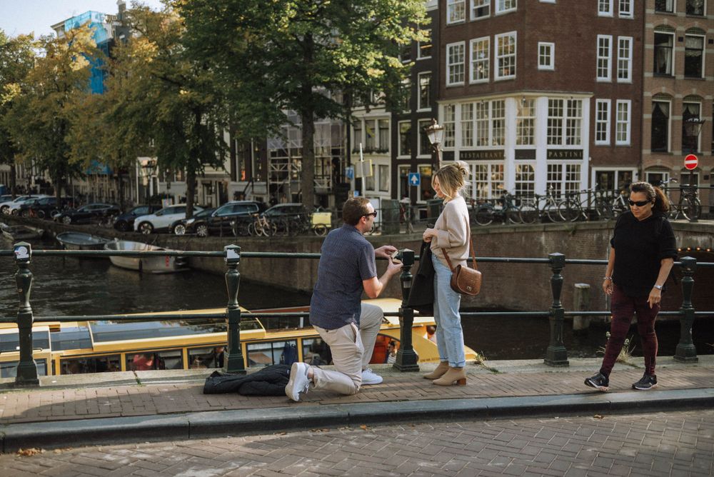 People enjoying sunny day by Amsterdam canal with boats and buildings.