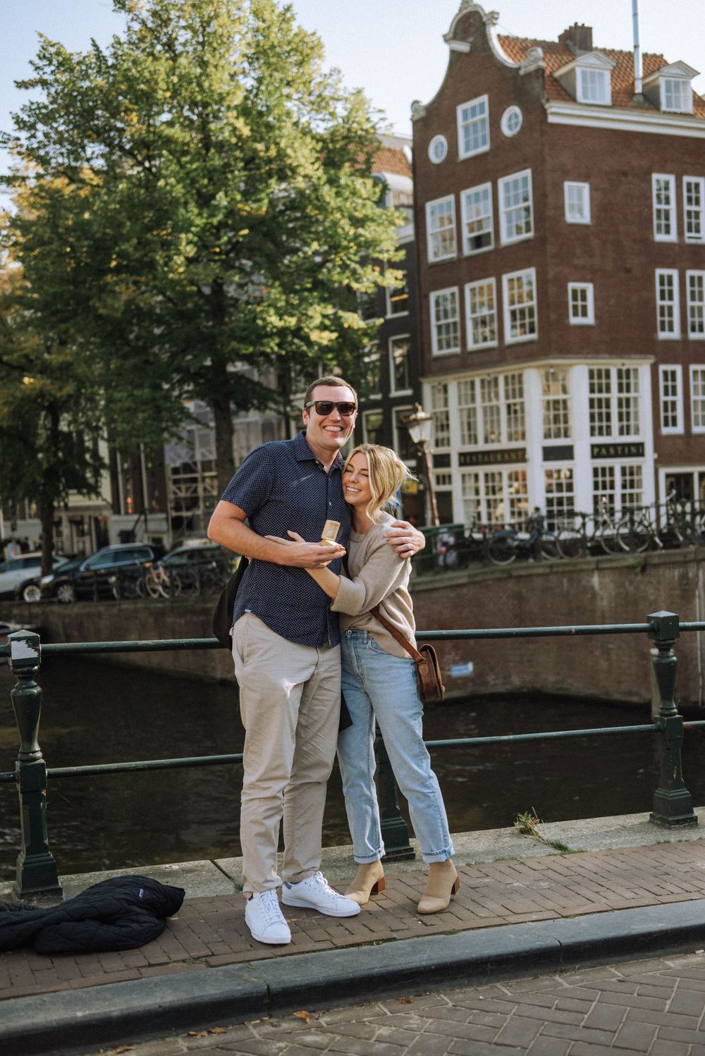A couple shares a romantic moment along a canal in Amsterdam with historic brick buildings in the background.