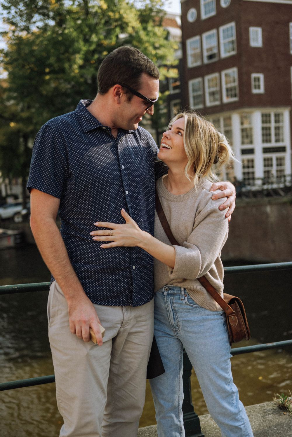 Couple embracing and laughing together by a canal with traditional Dutch architecture visible.