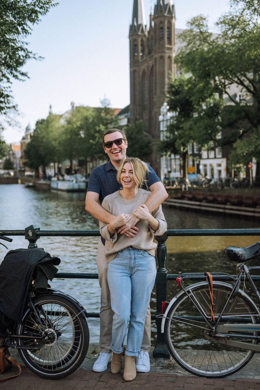 Couple posing playfully near parked bicycles along an Amsterdam canal with a church spire in the background.