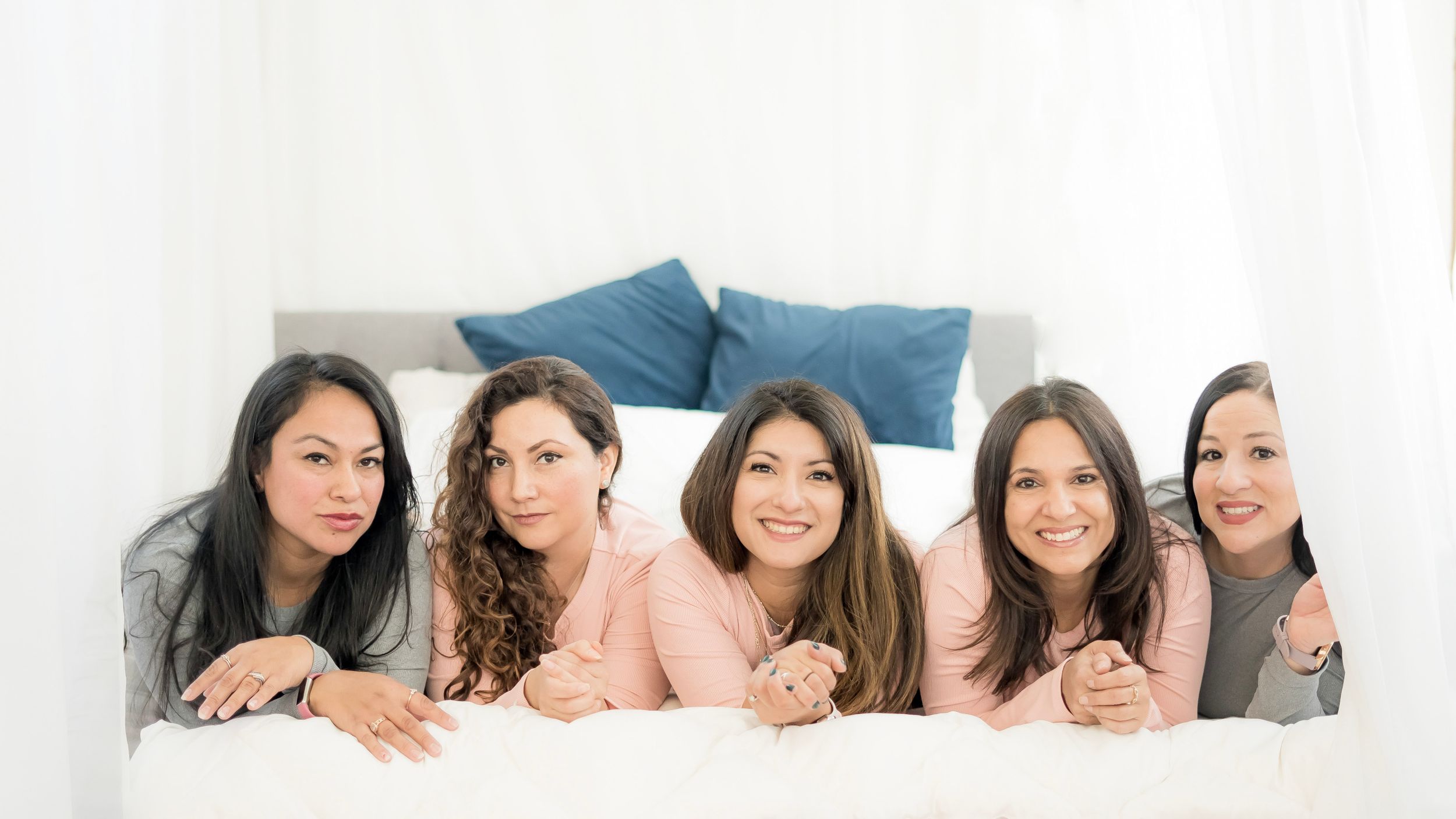 Five women friends lying on bed together in casual clothing.