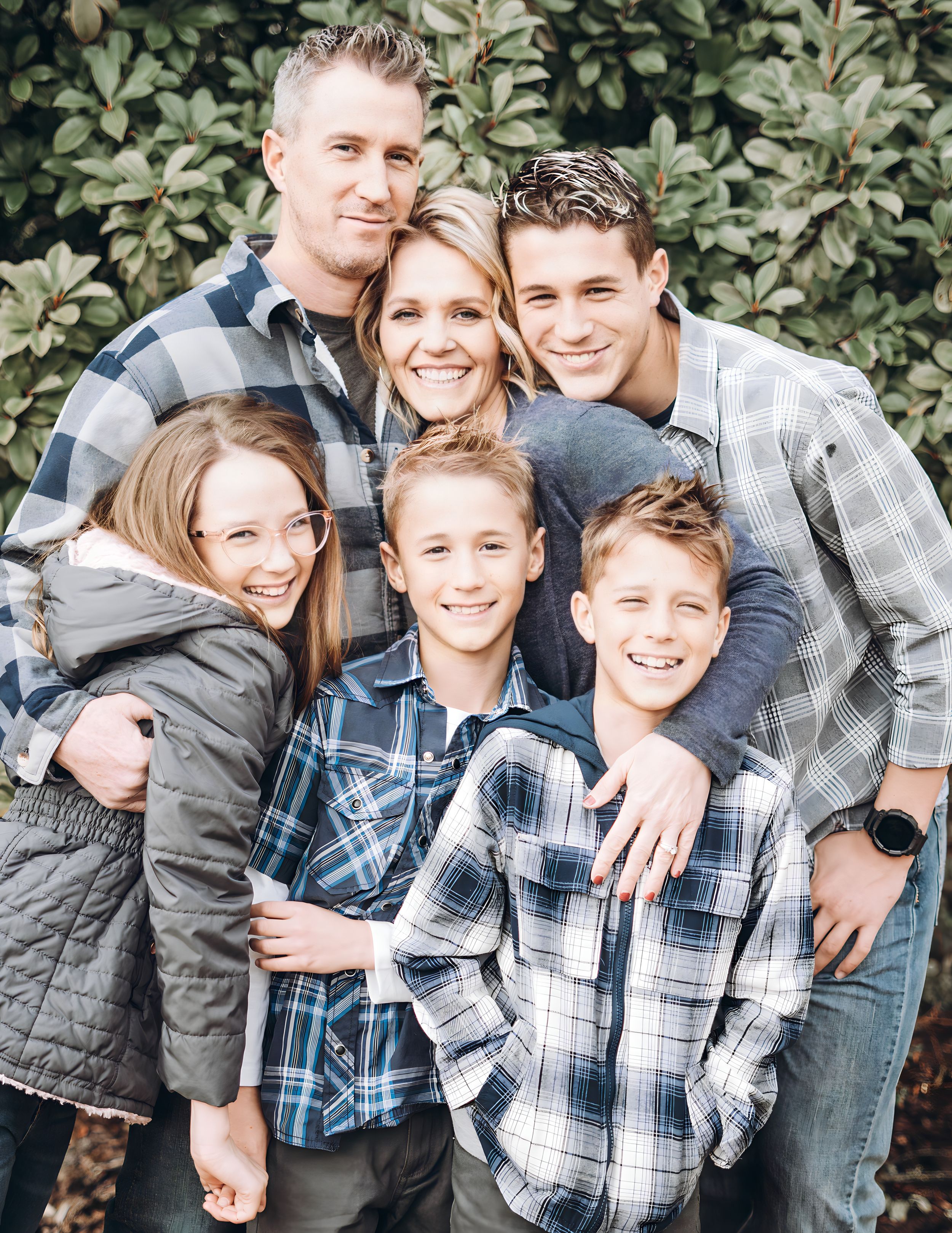 A family wearing coordinated plaid shirts poses together outdoors against greenery for a casual portrait.