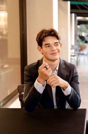 A well-dressed young man in a suit smiles while sitting at a dark table.