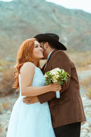 A romantic desert wedding photo of a couple embracing while holding a white bouquet against mountain backdrop.