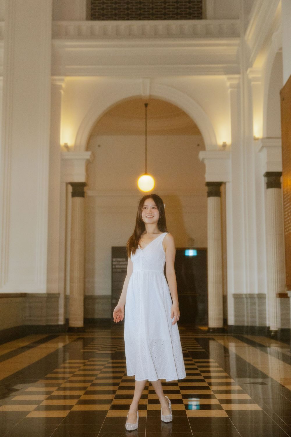 Figure in a white dress stands in an elegant hallway with marble floors and ornate archways.