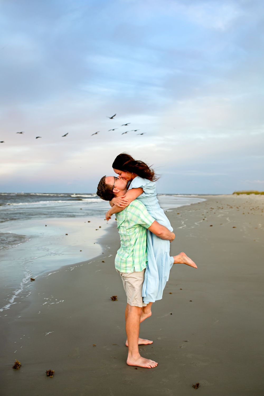 Playful beach moment with lifting embrace as birds fly overhead.