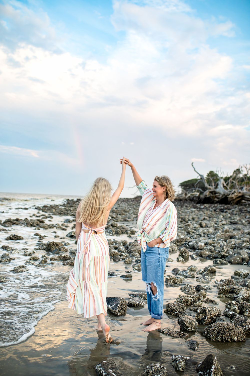 Two friends dancing together on rocky beach at sunset with rainbow in background.