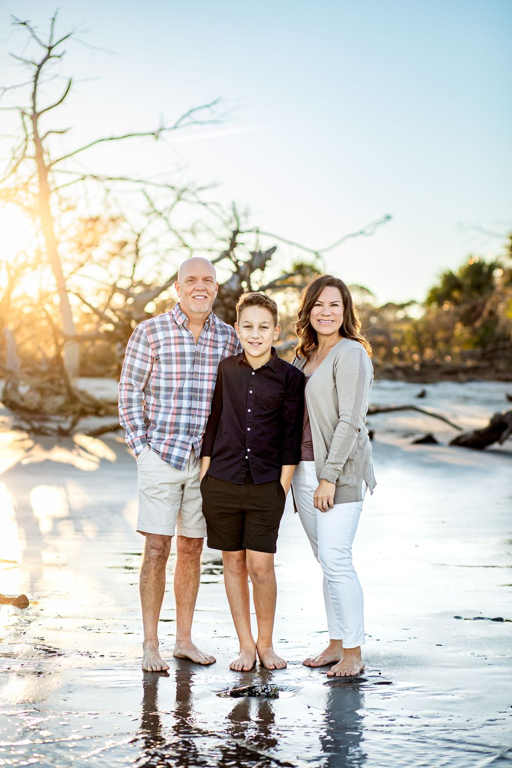 Family of three poses together on beach at sunset with driftwood backdrop.