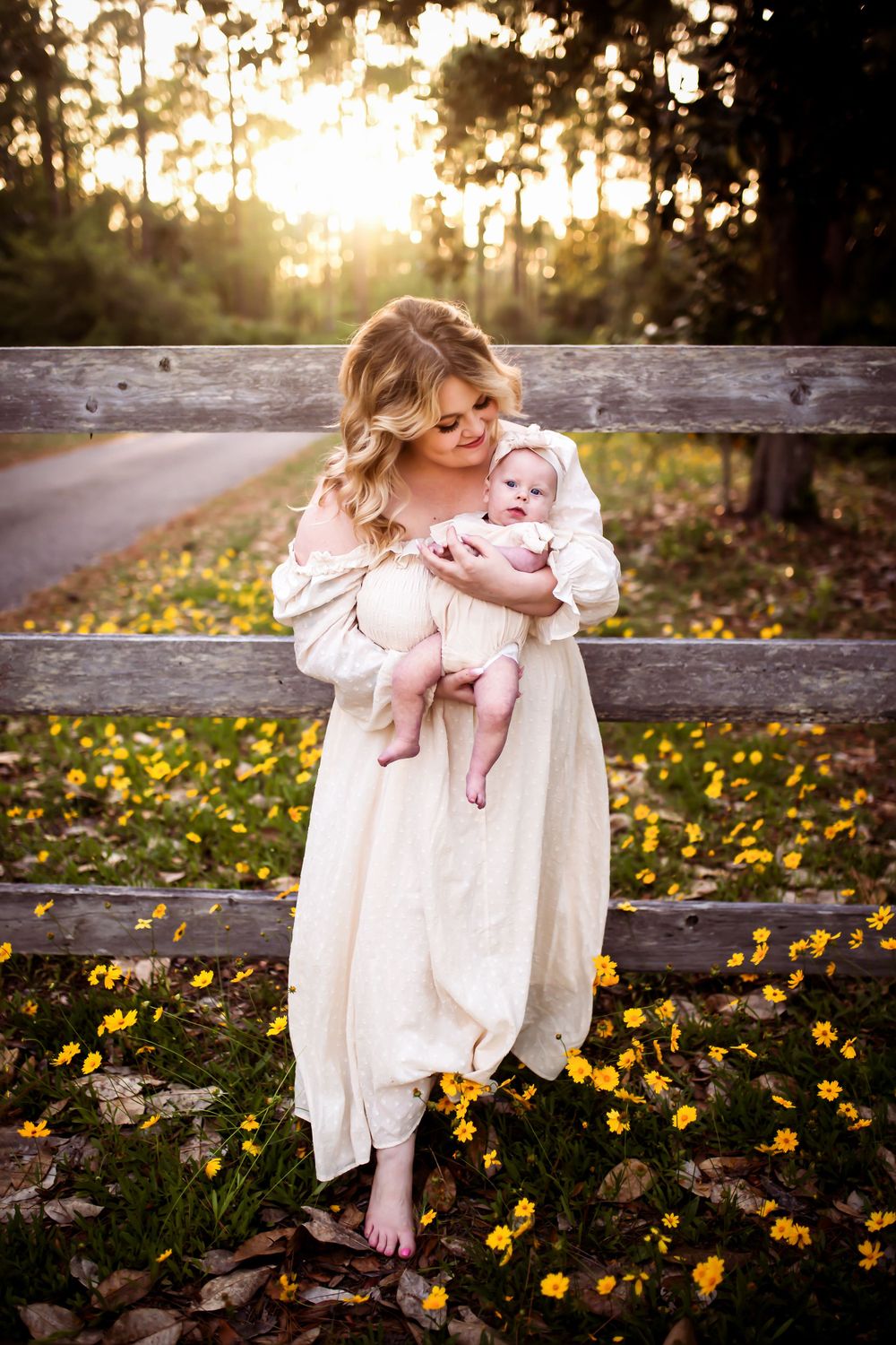 Mother and baby share tender moment by rustic fence surrounded by yellow wildflowers at sunset.