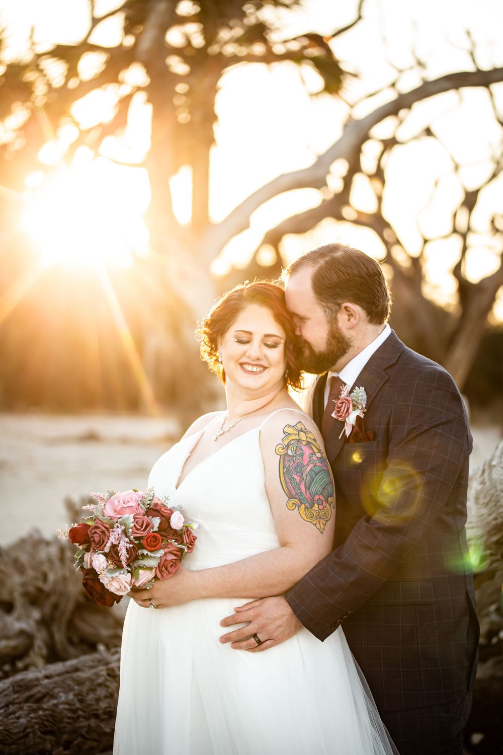 Romantic sunset beach wedding portrait with glowing sunburst through trees.
