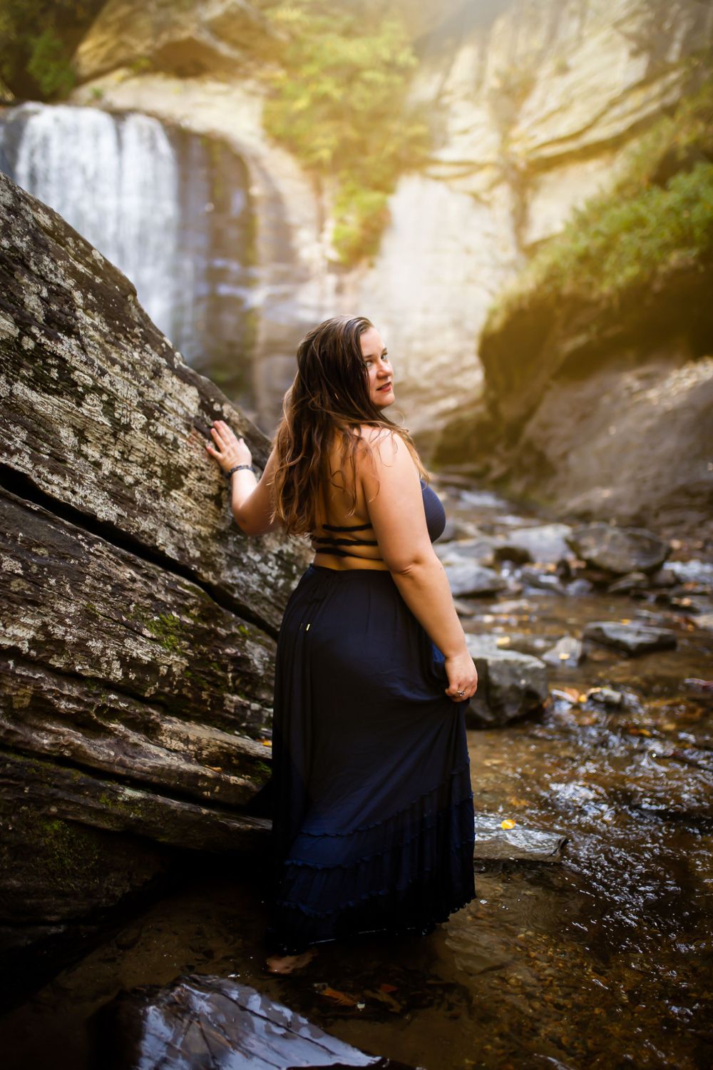 Person in dark dress poses near scenic waterfall during golden hour.