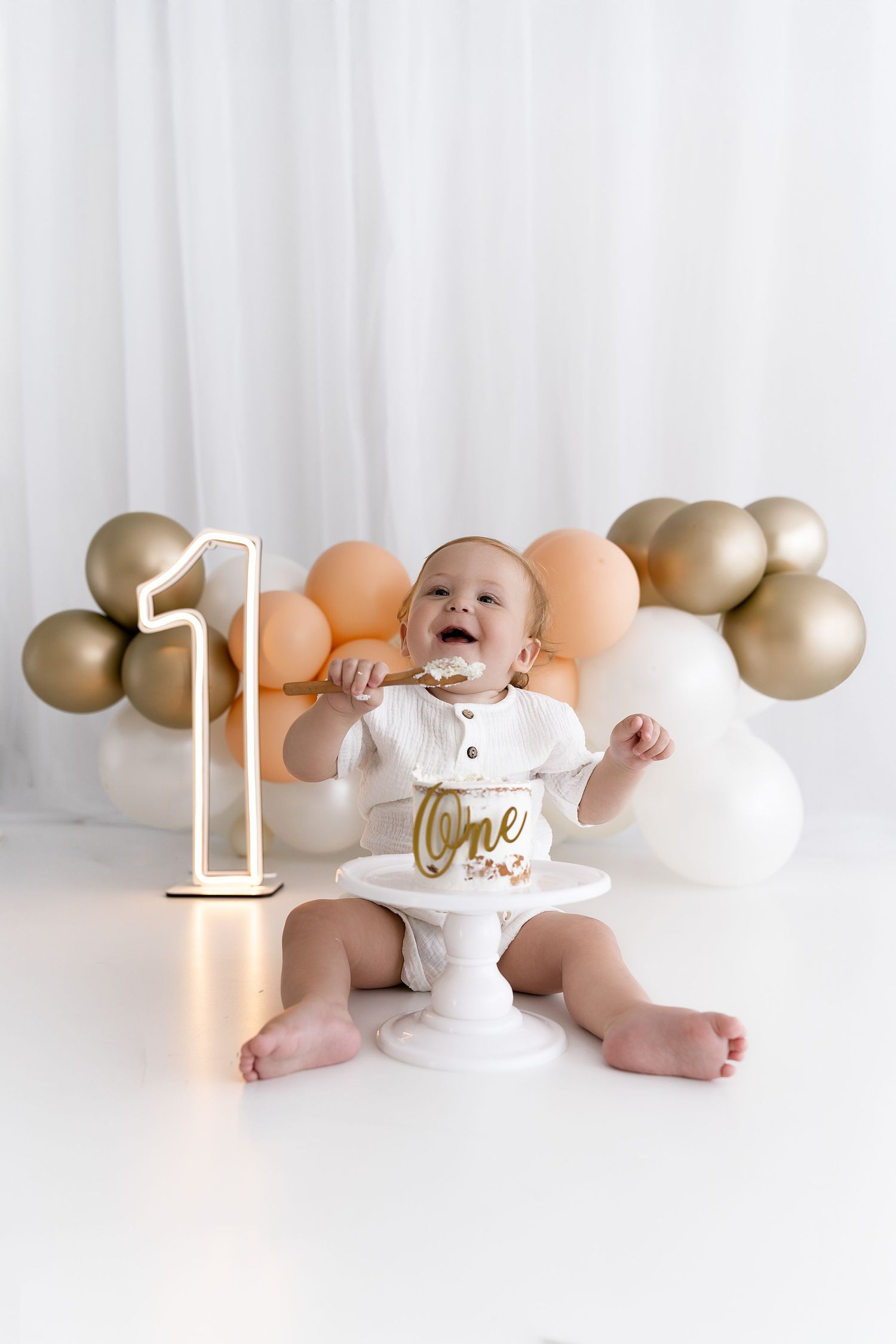 Baby celebrating first birthday with cake and gold balloon decorations.