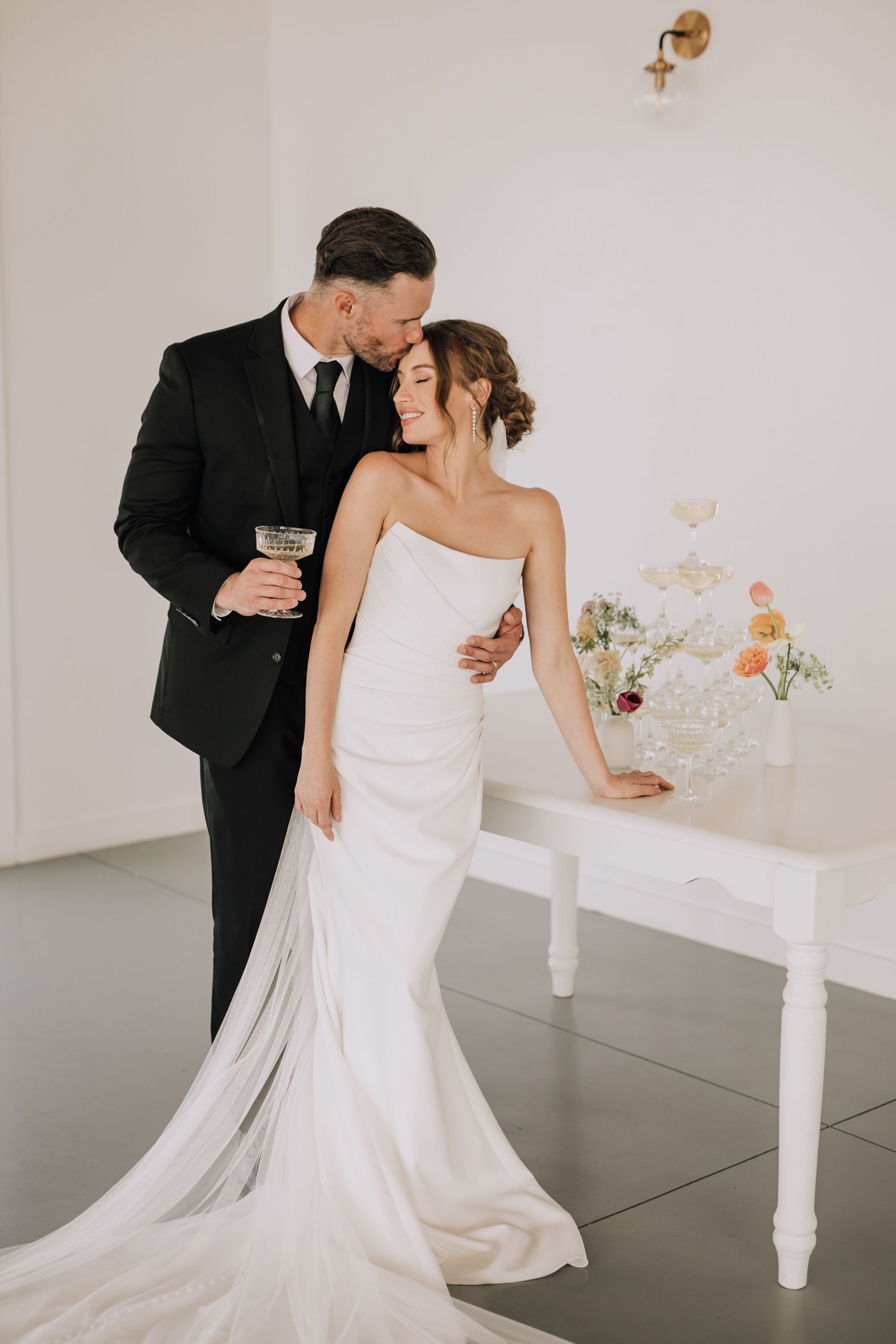 A romantic wedding scene with a couple embracing near a white table with floral arrangements in a minimalist setting.