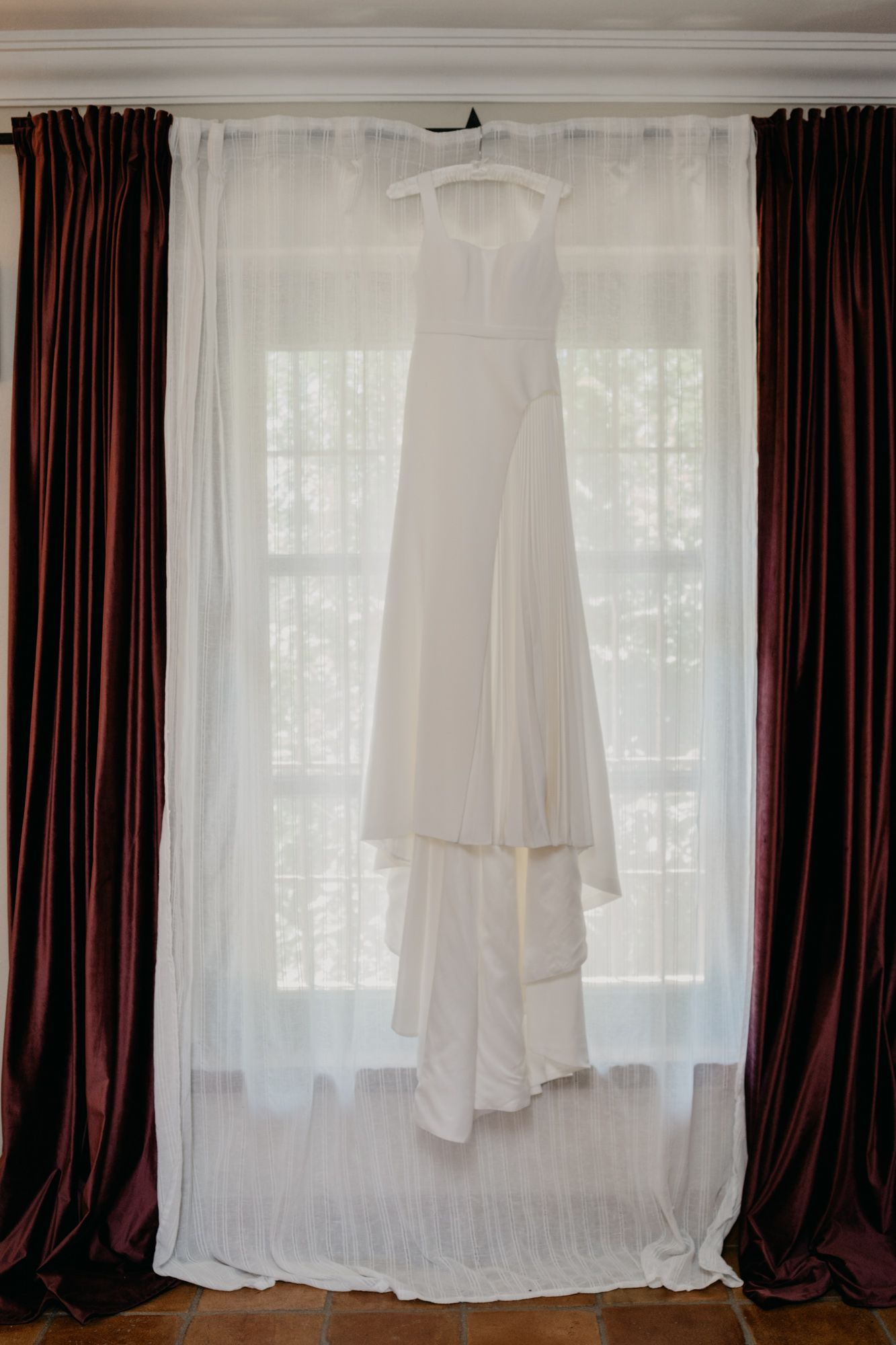 Elegant white wedding dress hanging in window between burgundy and white curtains.