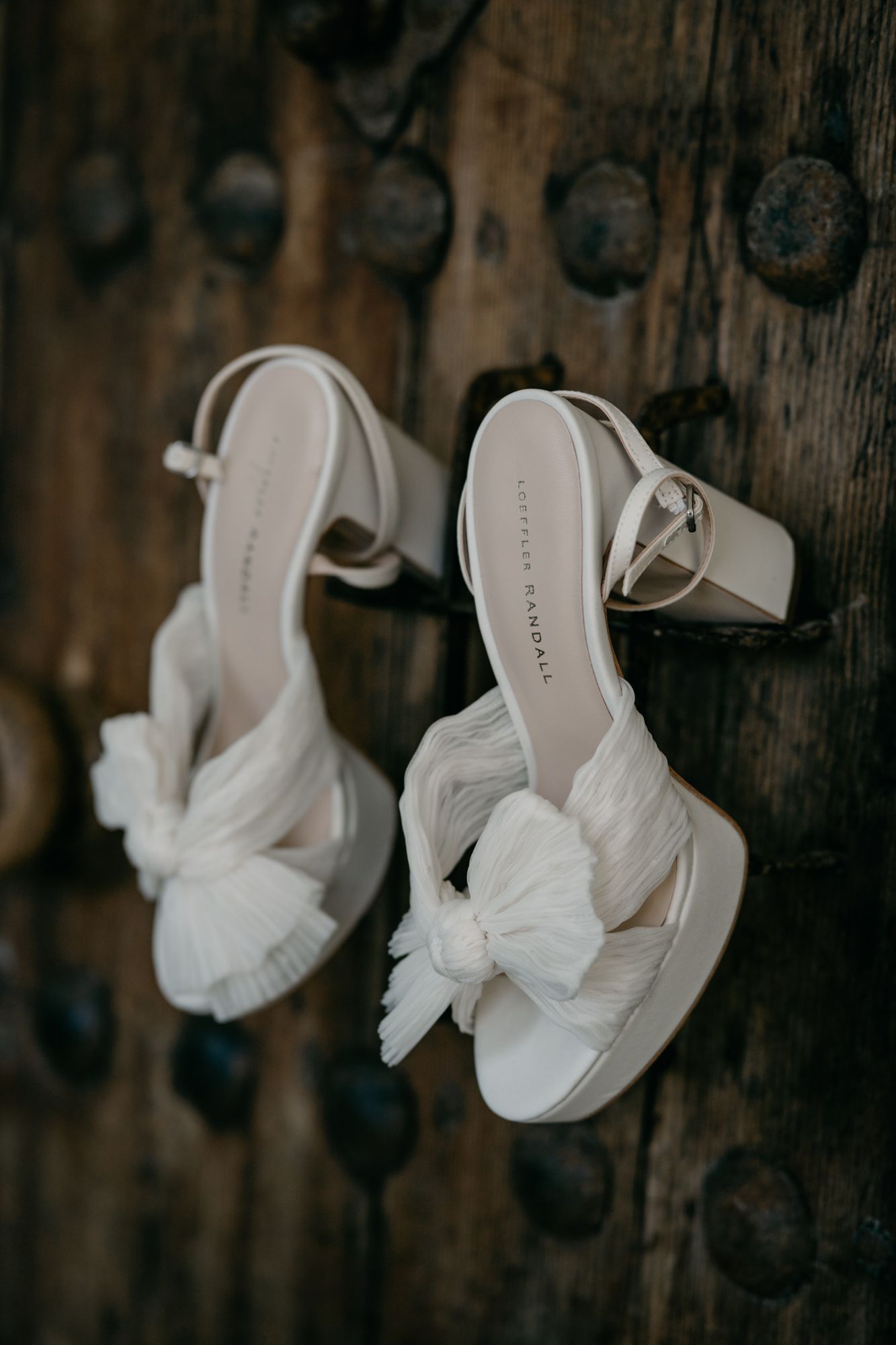 White bridal heels with bow detail photographed against rustic wooden door.