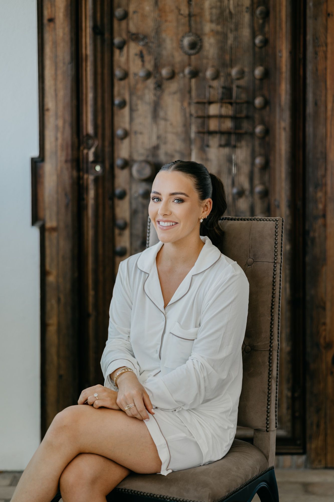 Woman in white robe sits smiling on upholstered chair against rustic wooden door.