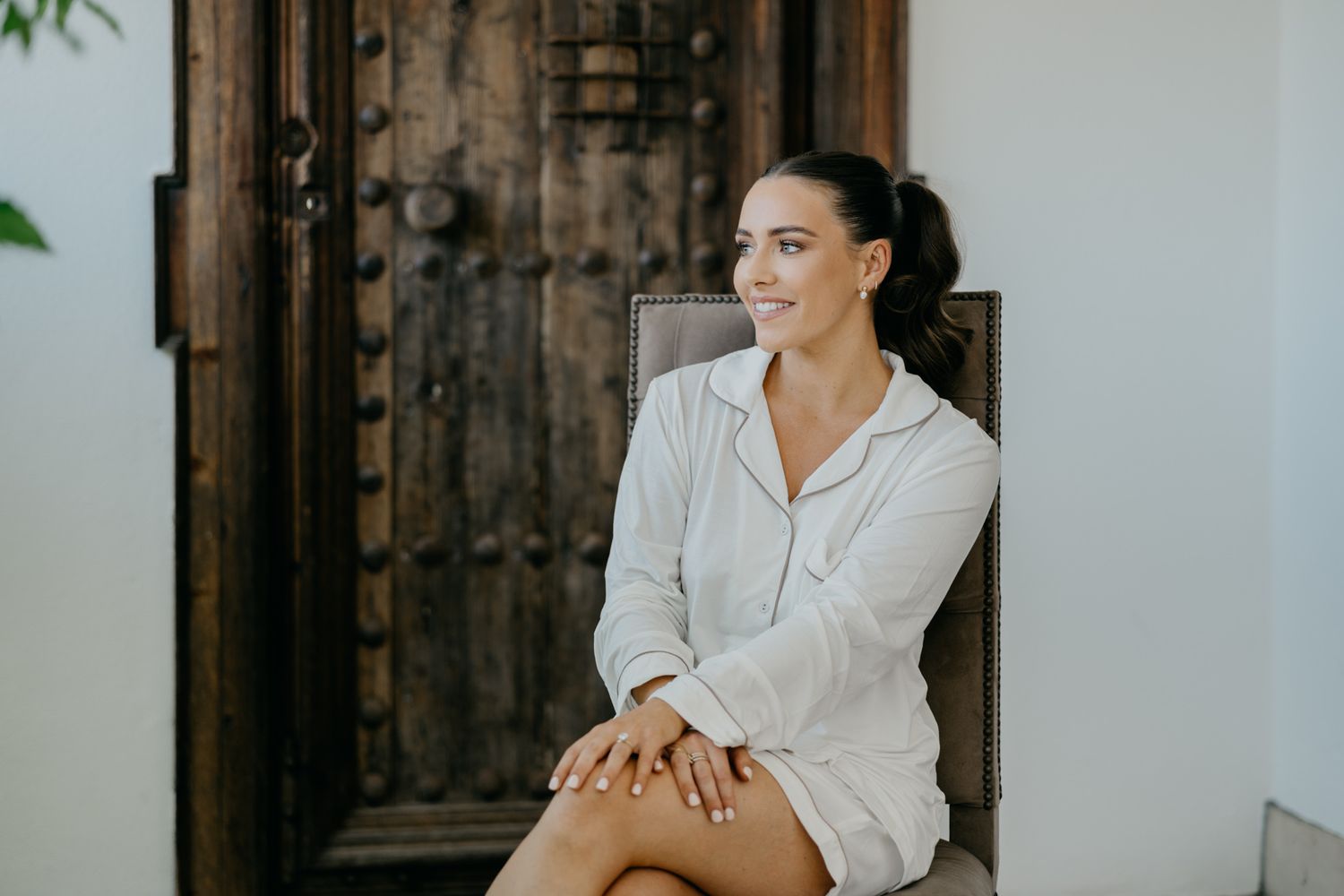 Woman in white robe sits casually on chair against vintage wooden door.