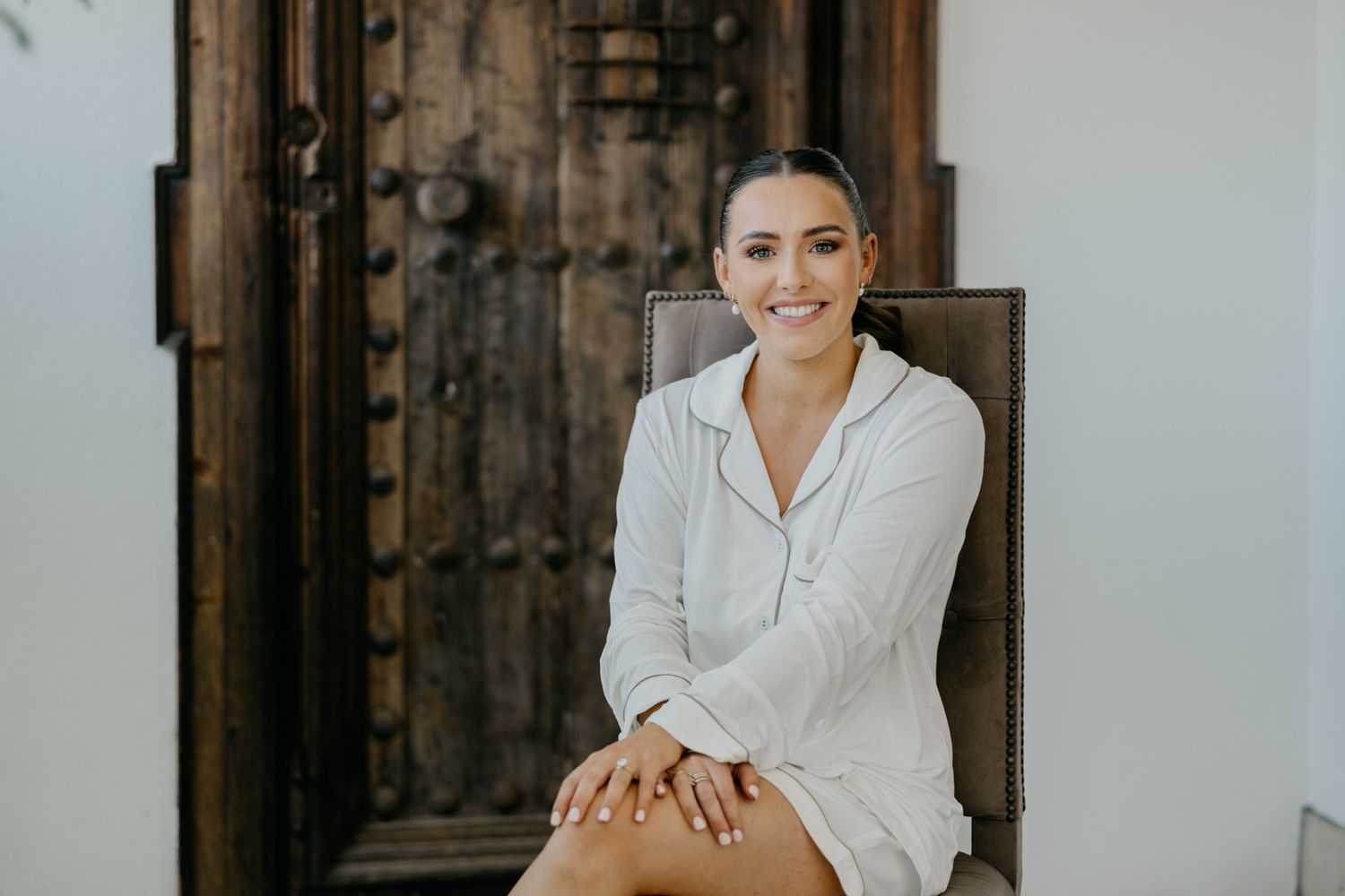 Woman in white robe poses on chair against rustic wooden backdrop.