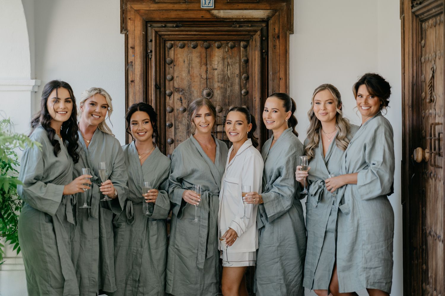 Bridal party in matching gray robes stands together smiling in white hallway.