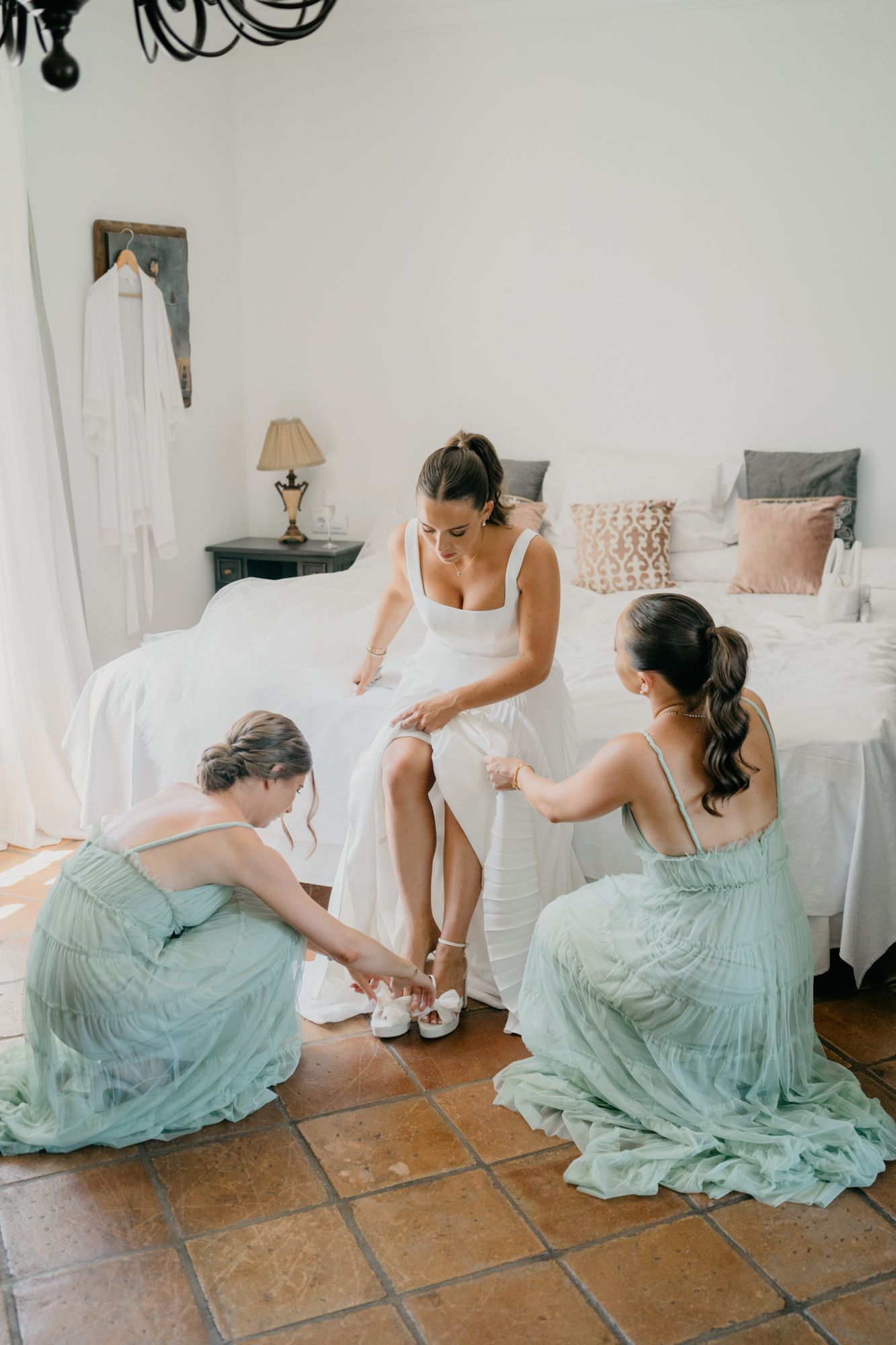 Bridesmaids in mint green dresses help with the bride's shoes during wedding preparations.