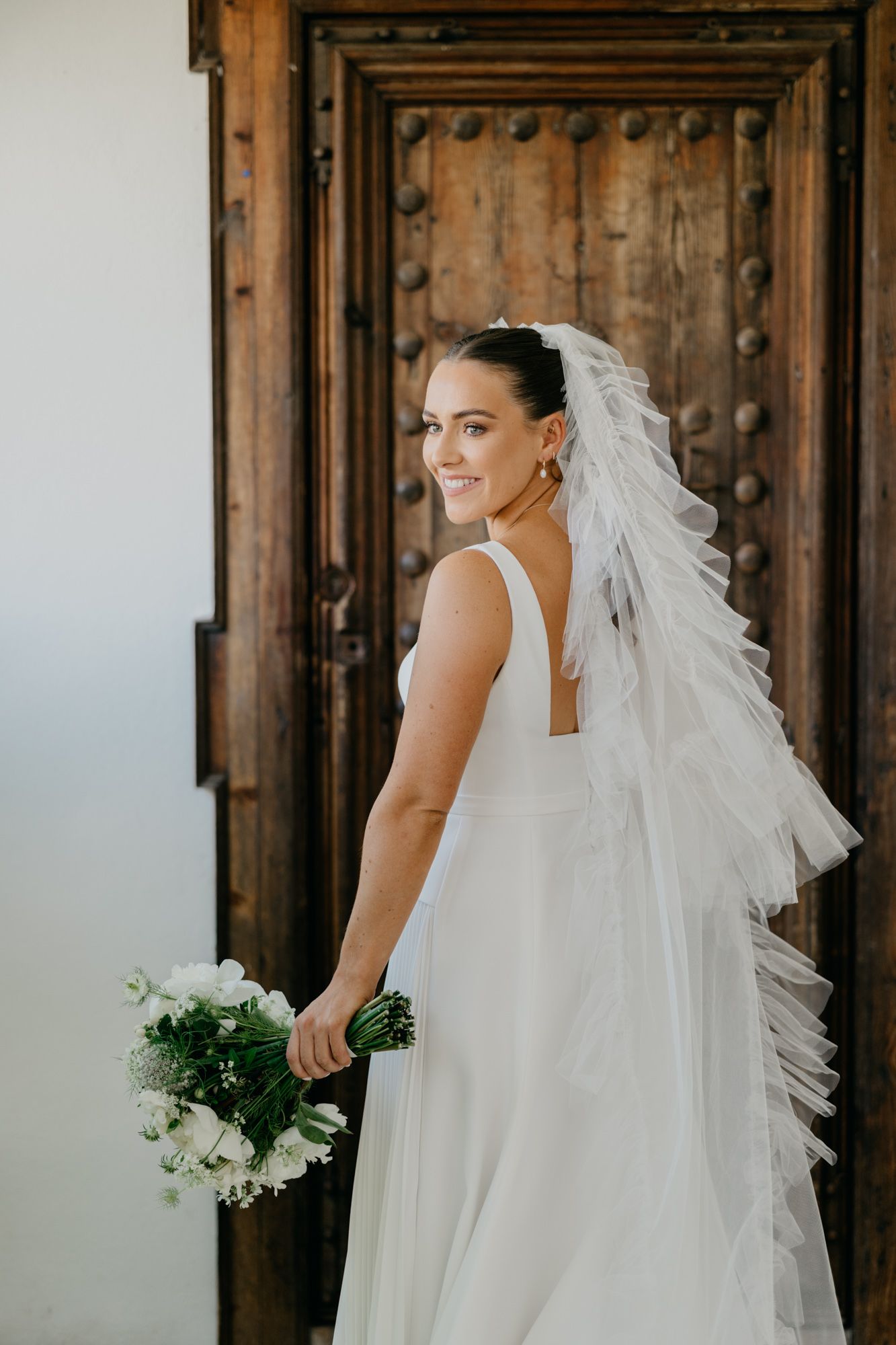 Bride in classic white dress and veil poses by rustic wooden door holding white bouquet.