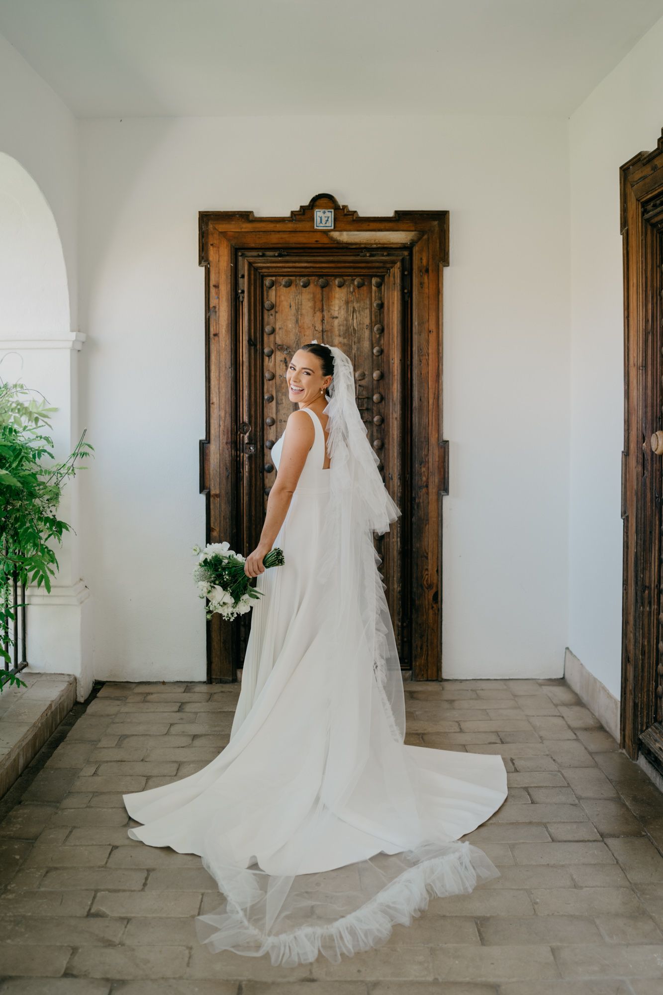 Full length view of bride in flowing white gown and veil standing by ornate wooden door.