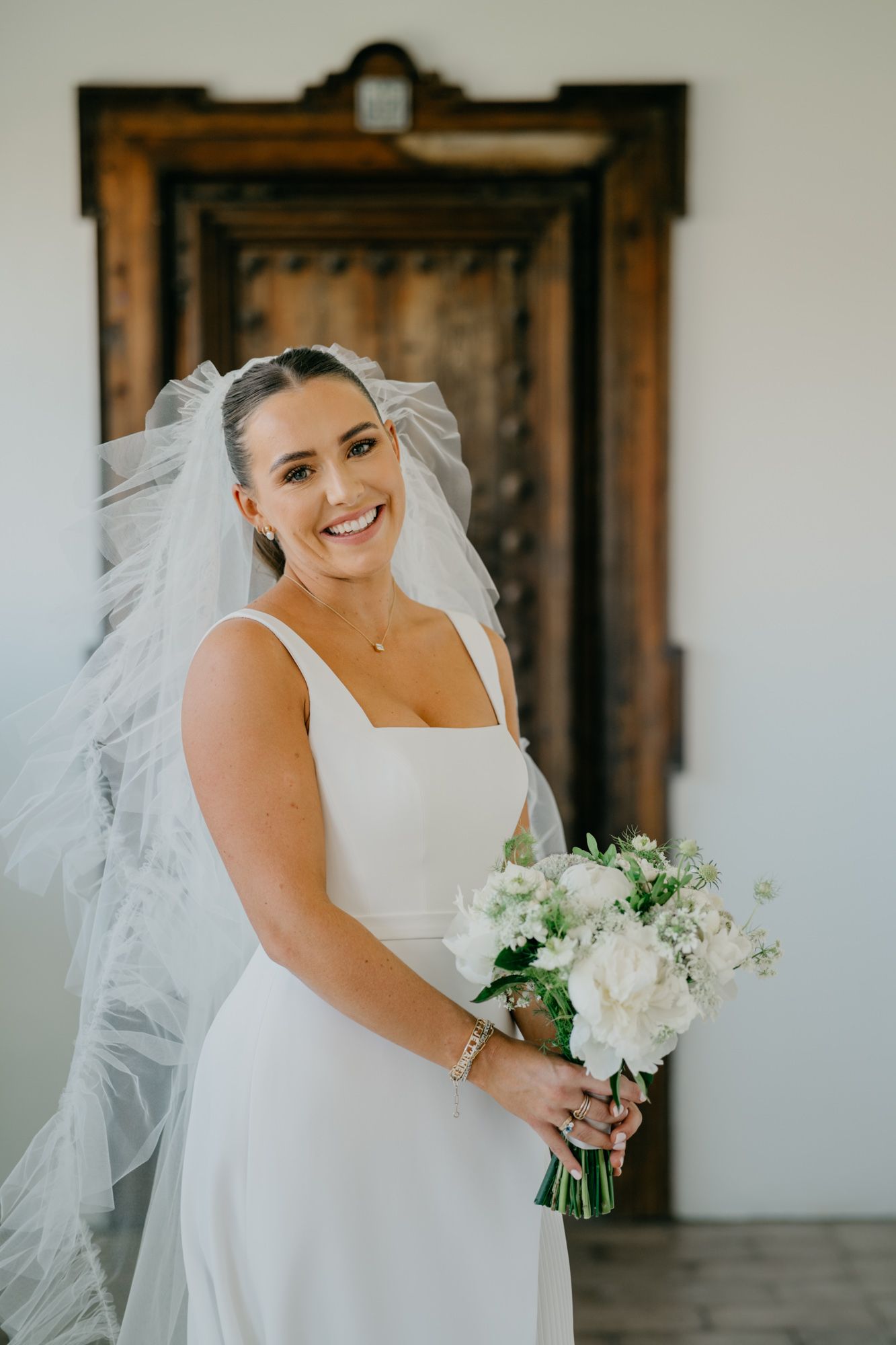 A bride in a classic white wedding dress and veil holds a white floral bouquet while standing in front of a wooden door.