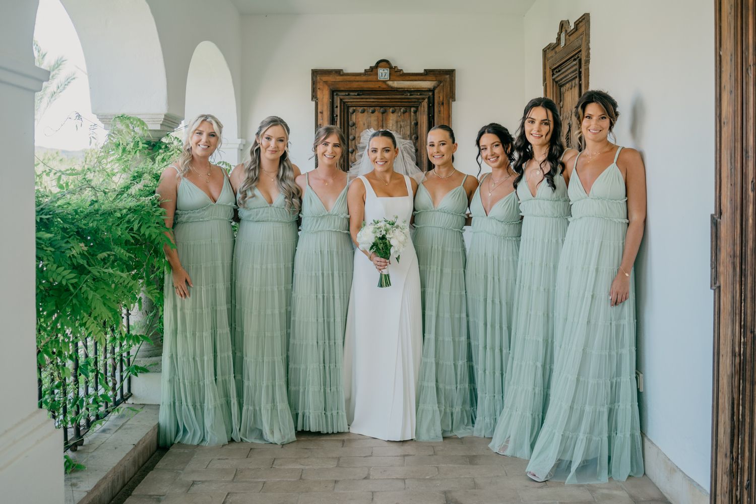 Bridal party wearing matching sage green dresses pose together on a white arched veranda.