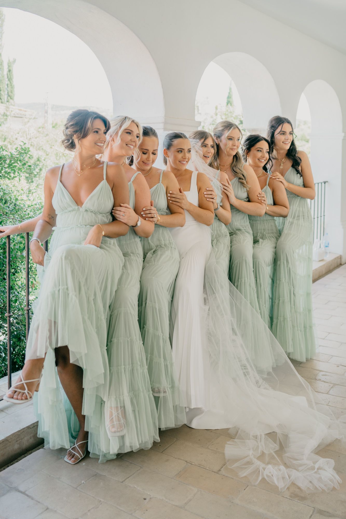 Bridesmaids in flowing mint green dresses laughing together with bride on a Mediterranean-style porch.