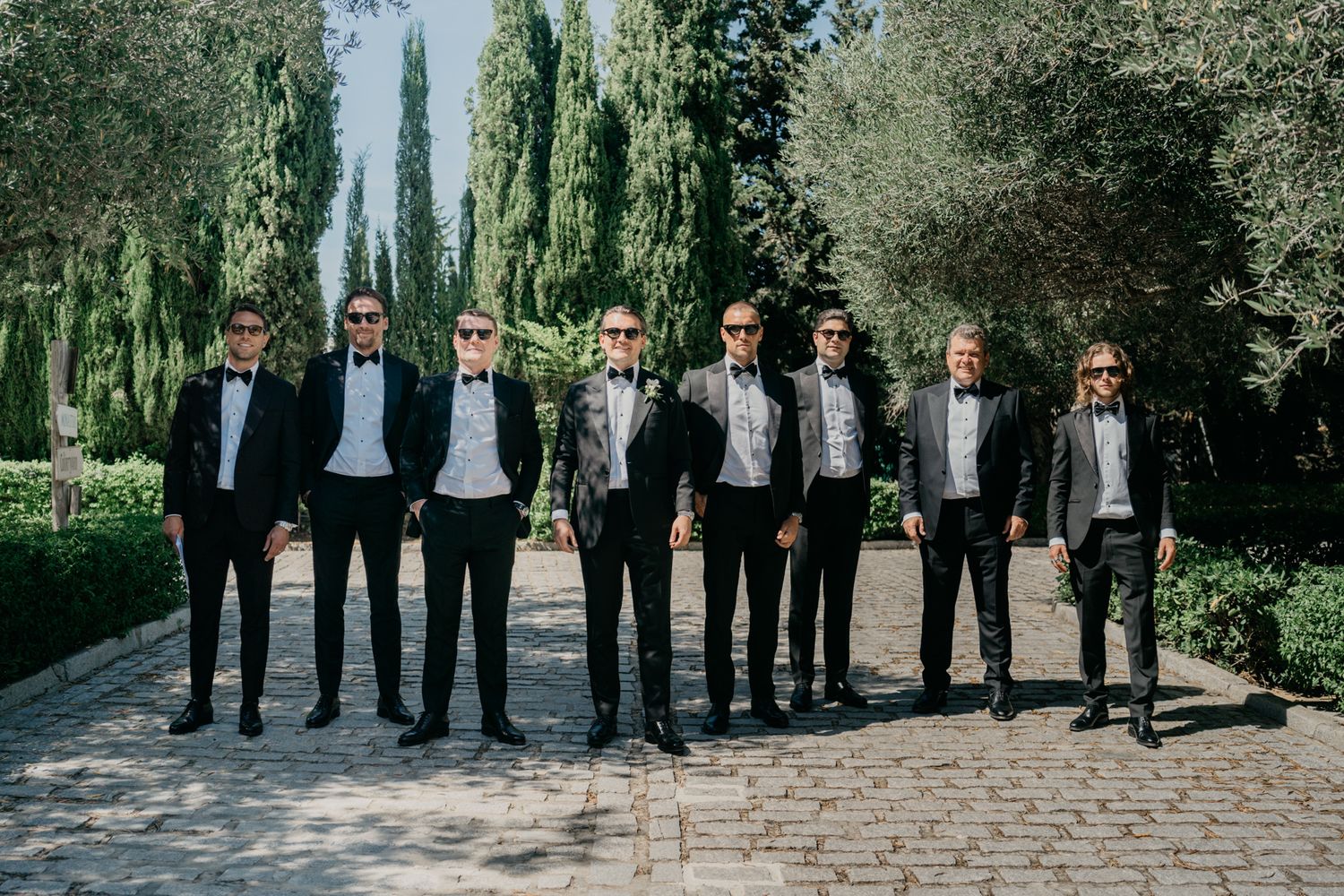 Groomsmen in black tuxedos stand in a line on a cypress tree-lined pathway.