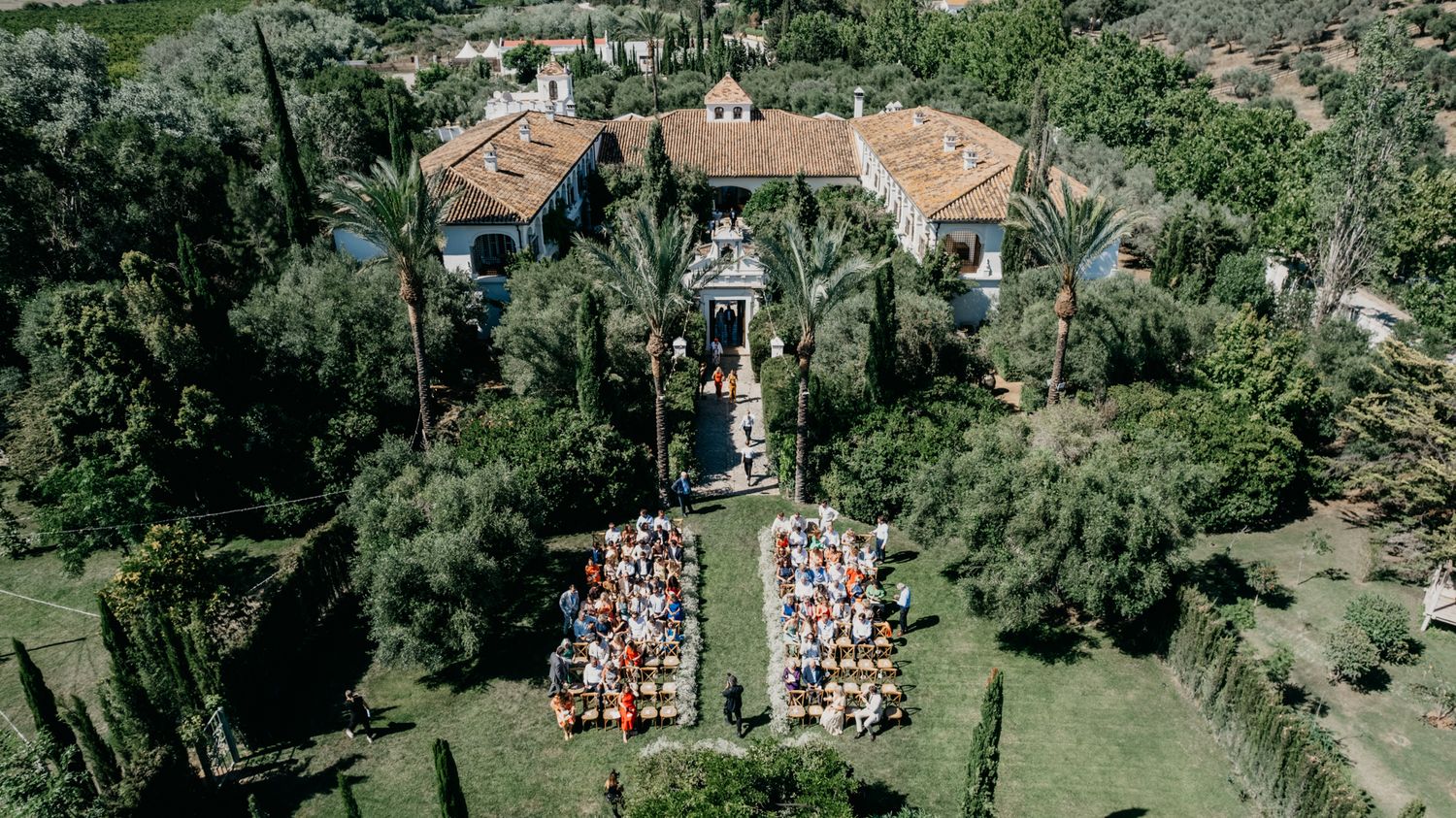 Drone shot of an outdoor wedding ceremony setup between two Mediterranean villa buildings.