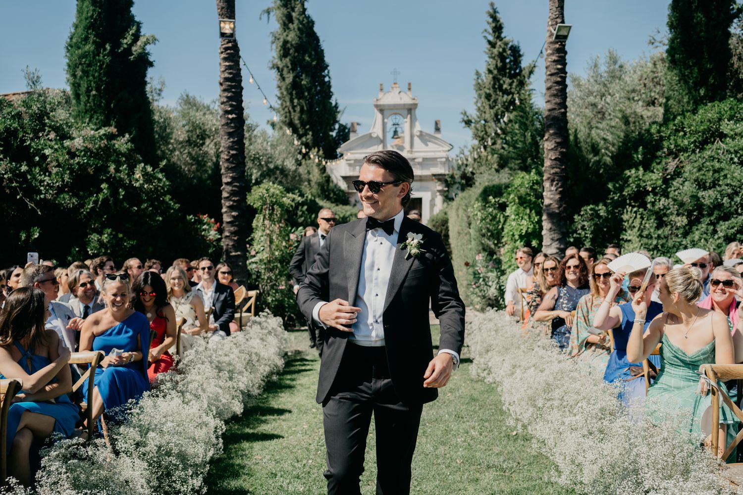 Wedding guests line the garden pathway during an outdoor ceremony with cypress trees and a white chapel in the background.