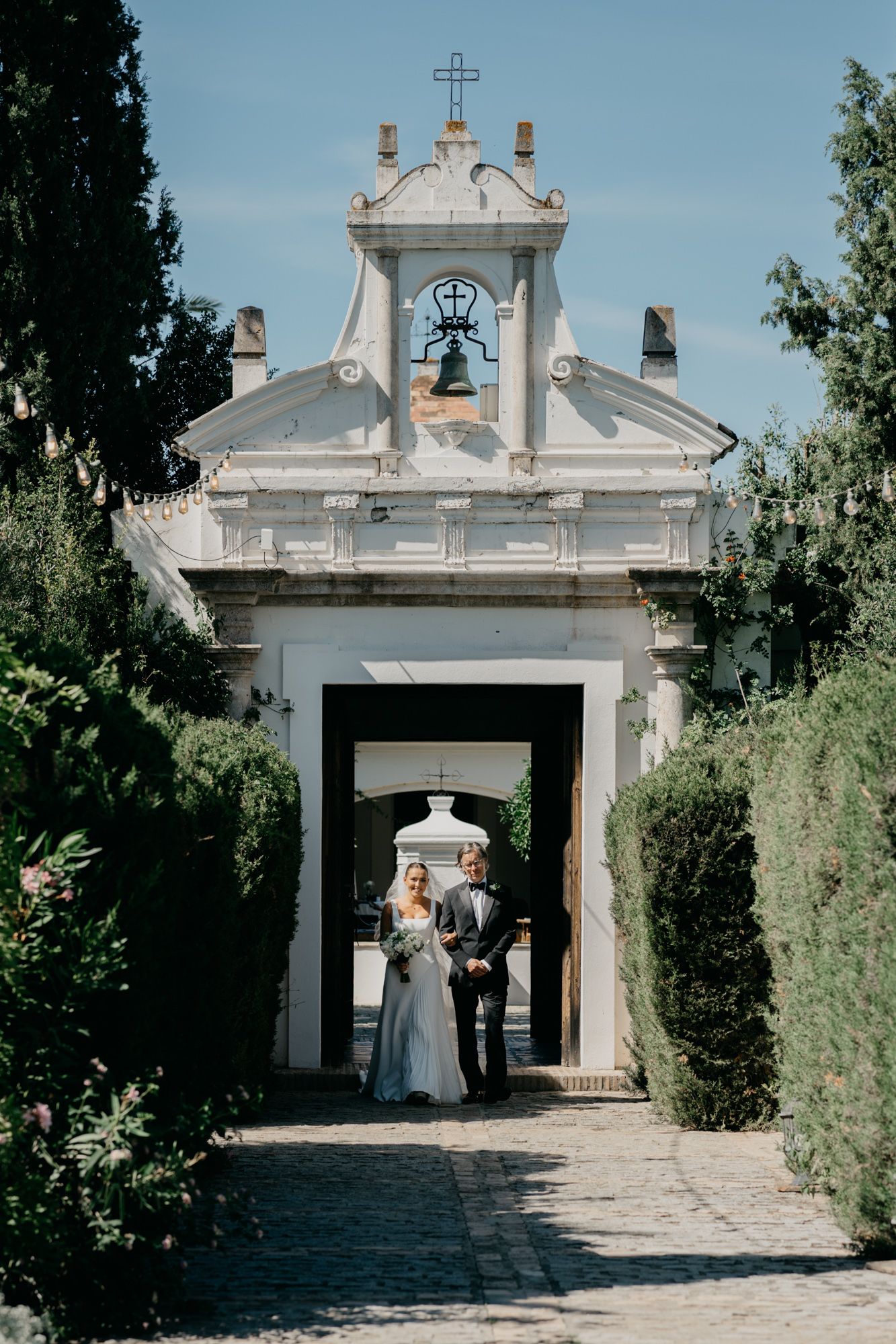 Historic white chapel entrance framed by cypress trees and stone pathways during a wedding ceremony.
