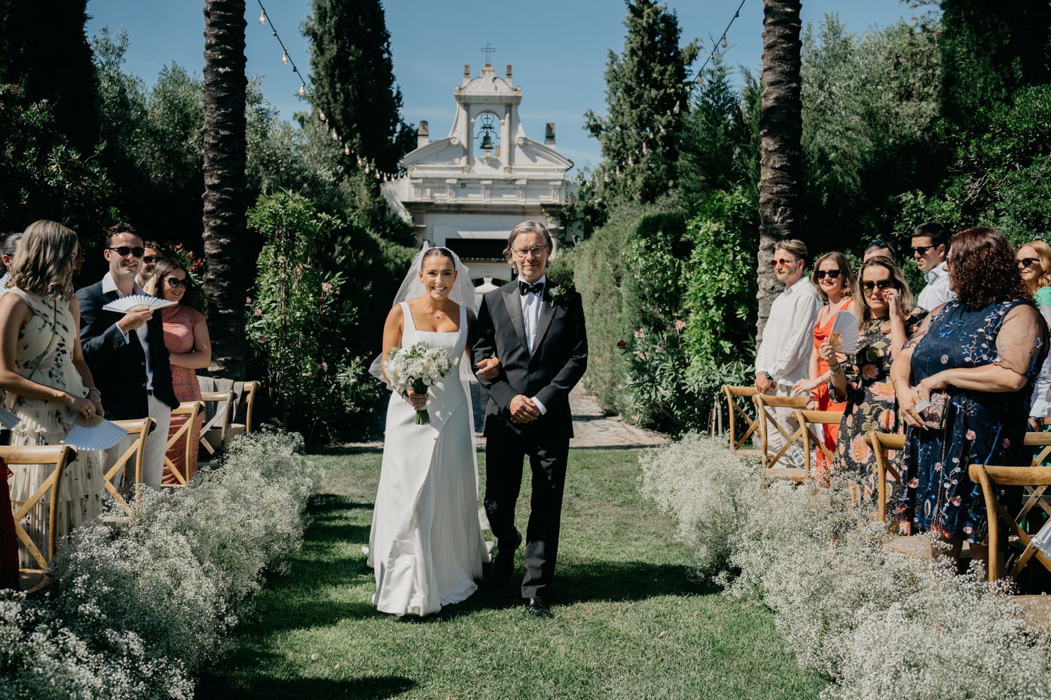 Garden wedding ceremony with palm trees and white chapel backdrop as guests watch from wooden chairs.