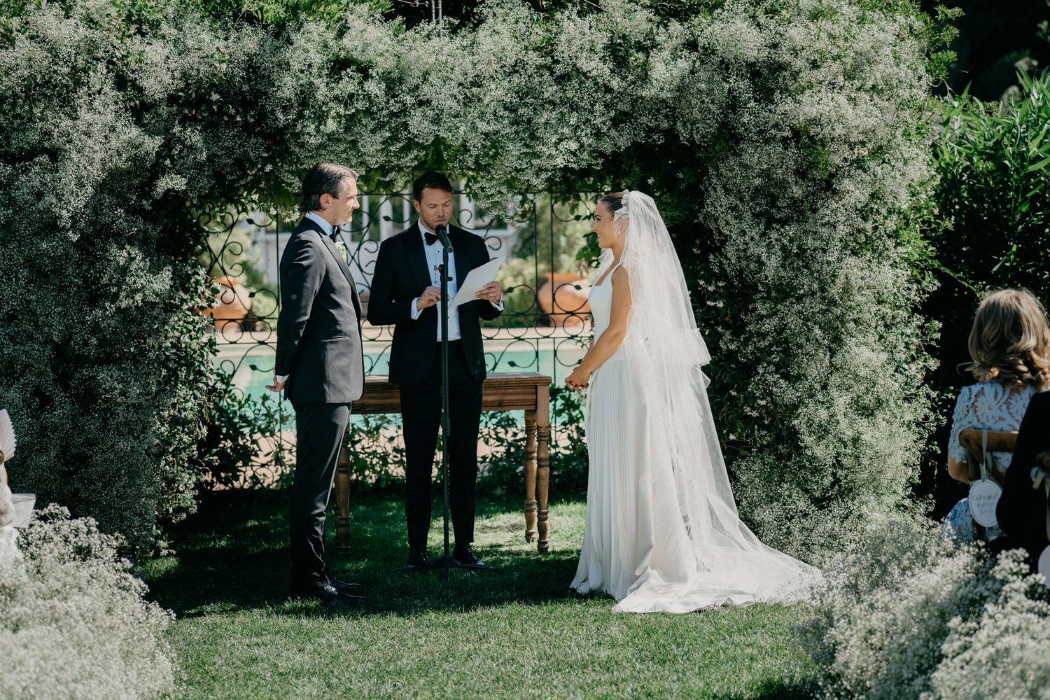 Outdoor wedding ceremony under flowering archway with guests seated on lawn.