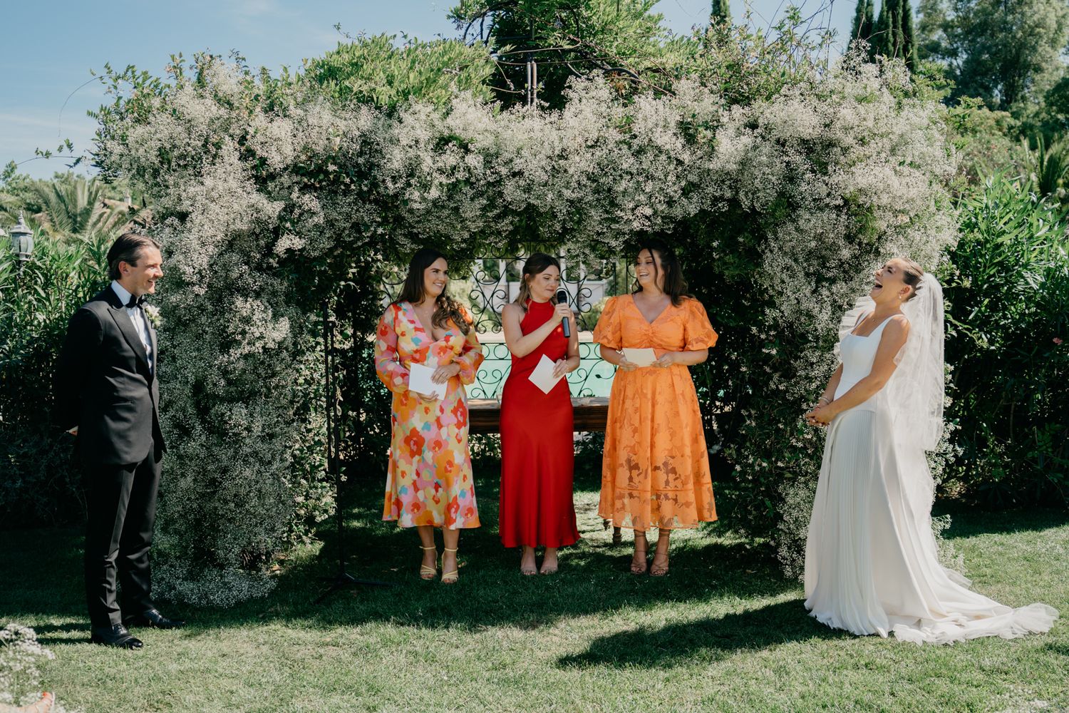 Wedding party poses under floral arch during garden ceremony with colorful attire.