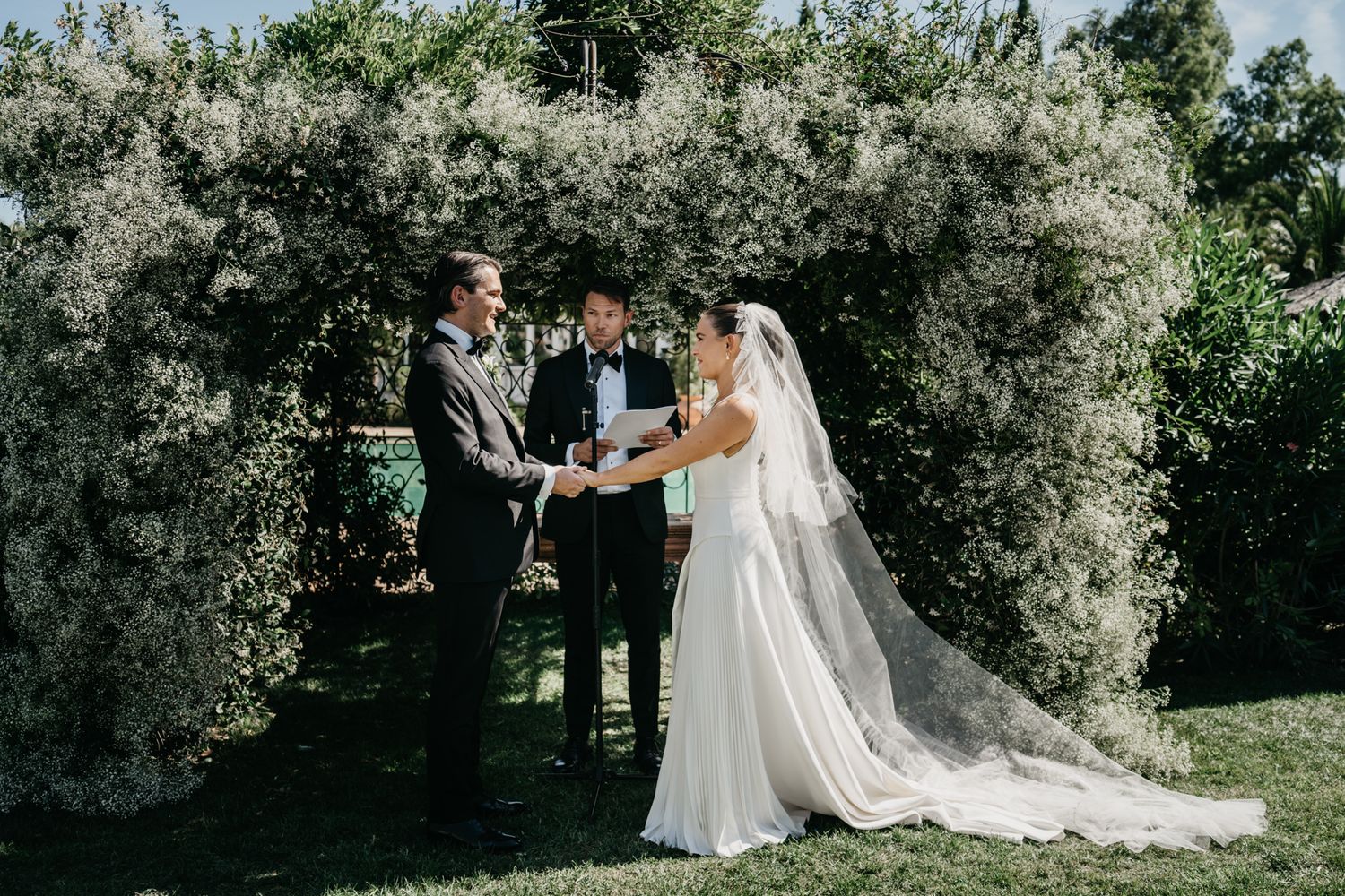 A wedding ceremony takes place under a beautiful floral arch in an outdoor garden setting.