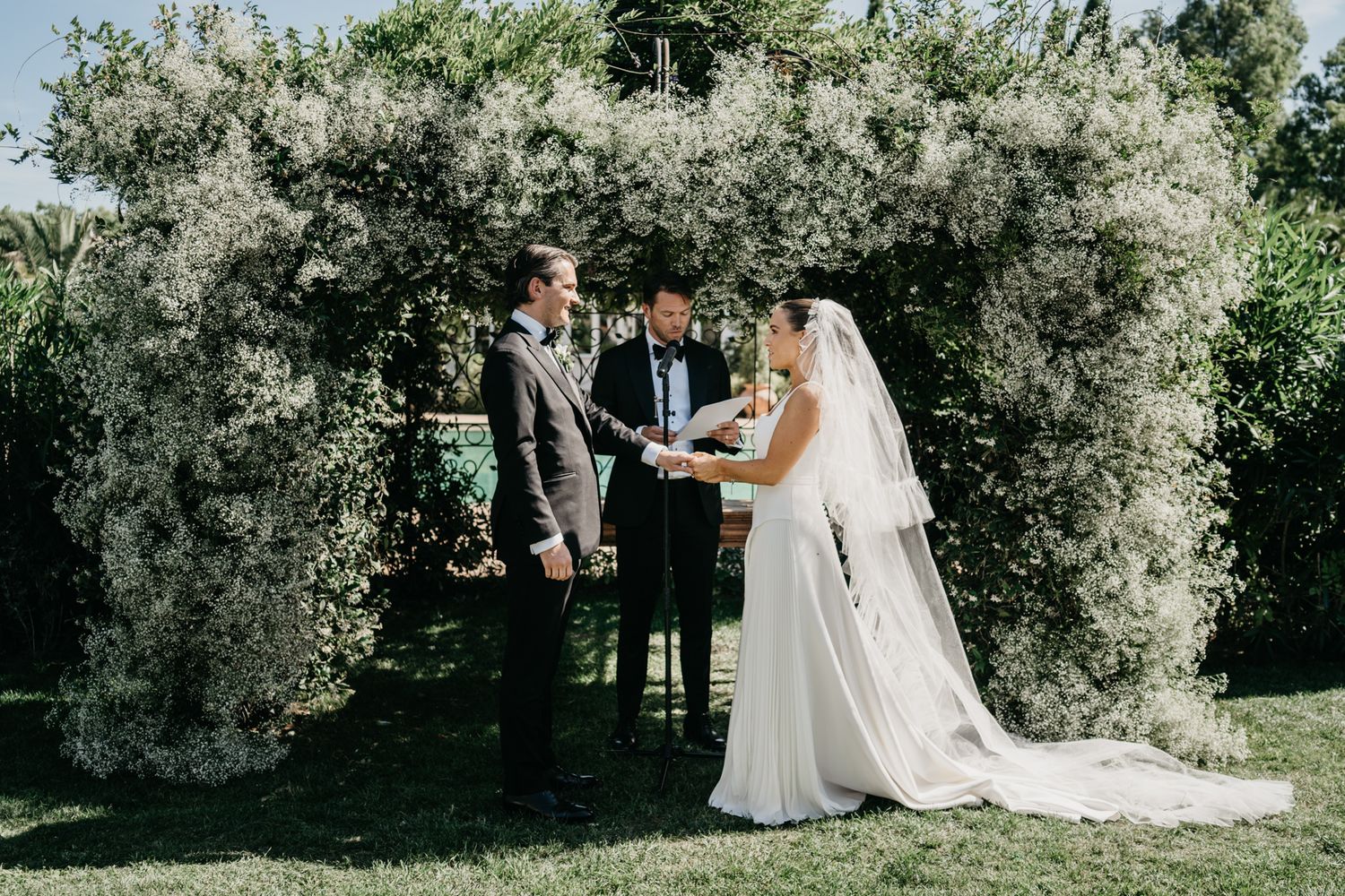 An intimate wedding ceremony beneath a lush floral archway in a sunlit garden.